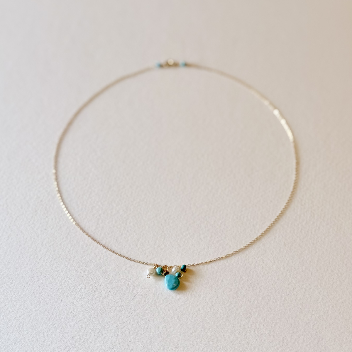 A delicate gold chain necklace with a small cluster of turquoise blue beads and a central turquoise stone pendant, displayed on a textured off-white surface.