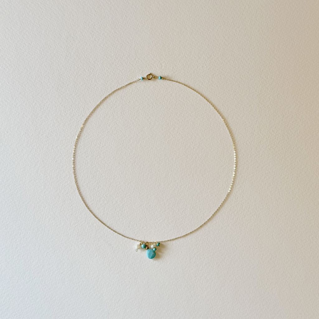 A delicate gold chain necklace with a small cluster of turquoise, white, and gold beads at the center, displayed against a plain off-white wall.