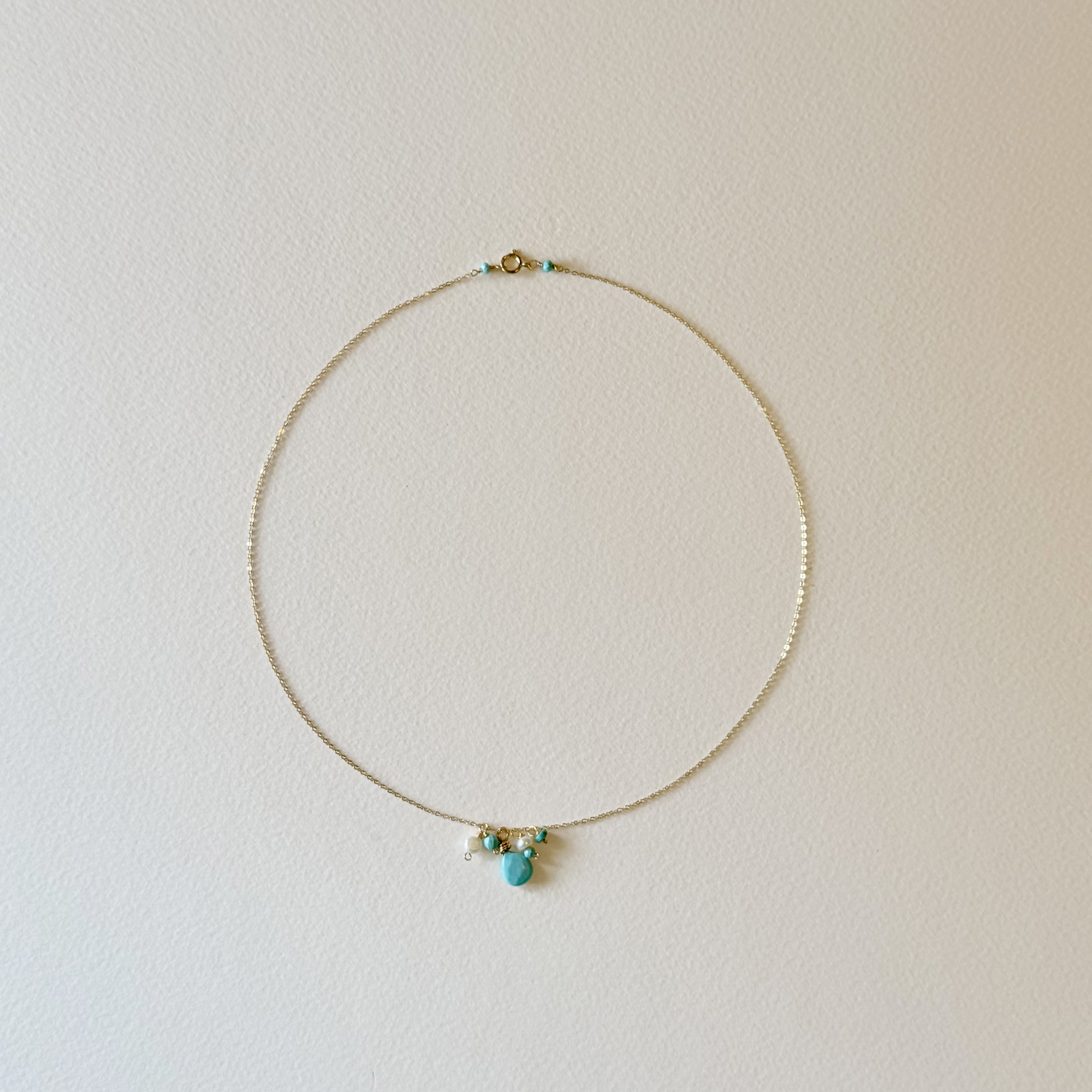 A delicate gold chain necklace with a small cluster of turquoise, white, and gold beads at the center, displayed against a plain off-white wall.