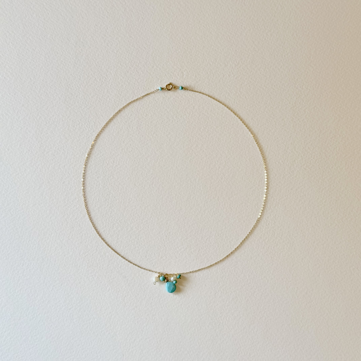 A delicate gold chain necklace with a small cluster of turquoise, white, and gold beads at the center, displayed against a plain off-white wall.