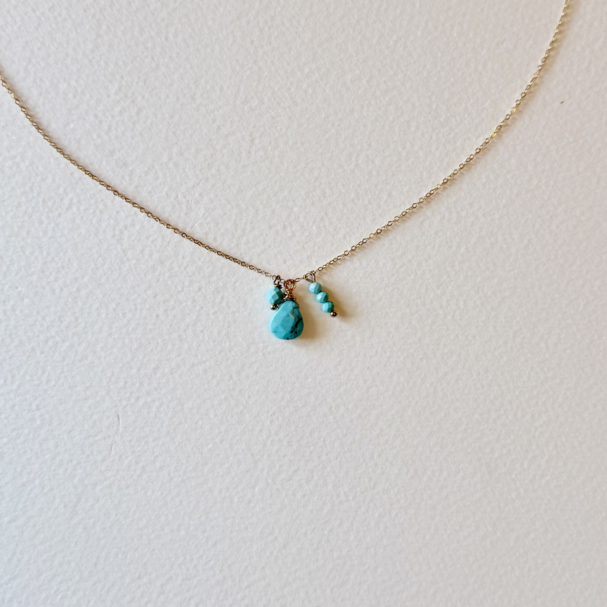 A delicate gold chain necklace featuring a small turquoise stone pendant and two tiny turquoise bead charms, displayed on a textured off-white background.