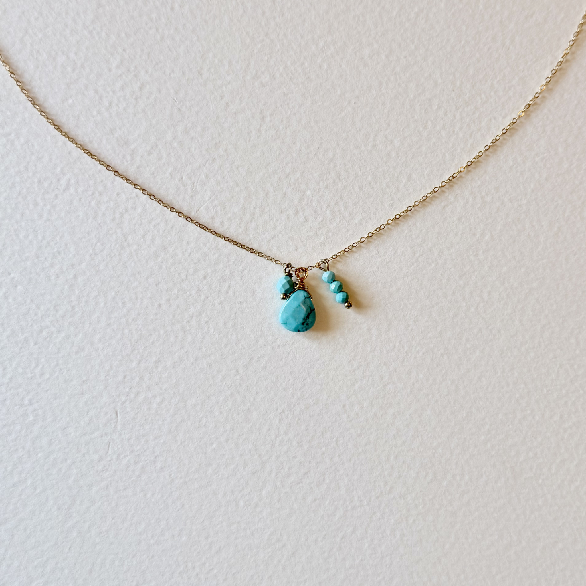 A delicate gold chain necklace featuring a central cluster of small turquoise-colored stones, displayed on a textured off-white background.