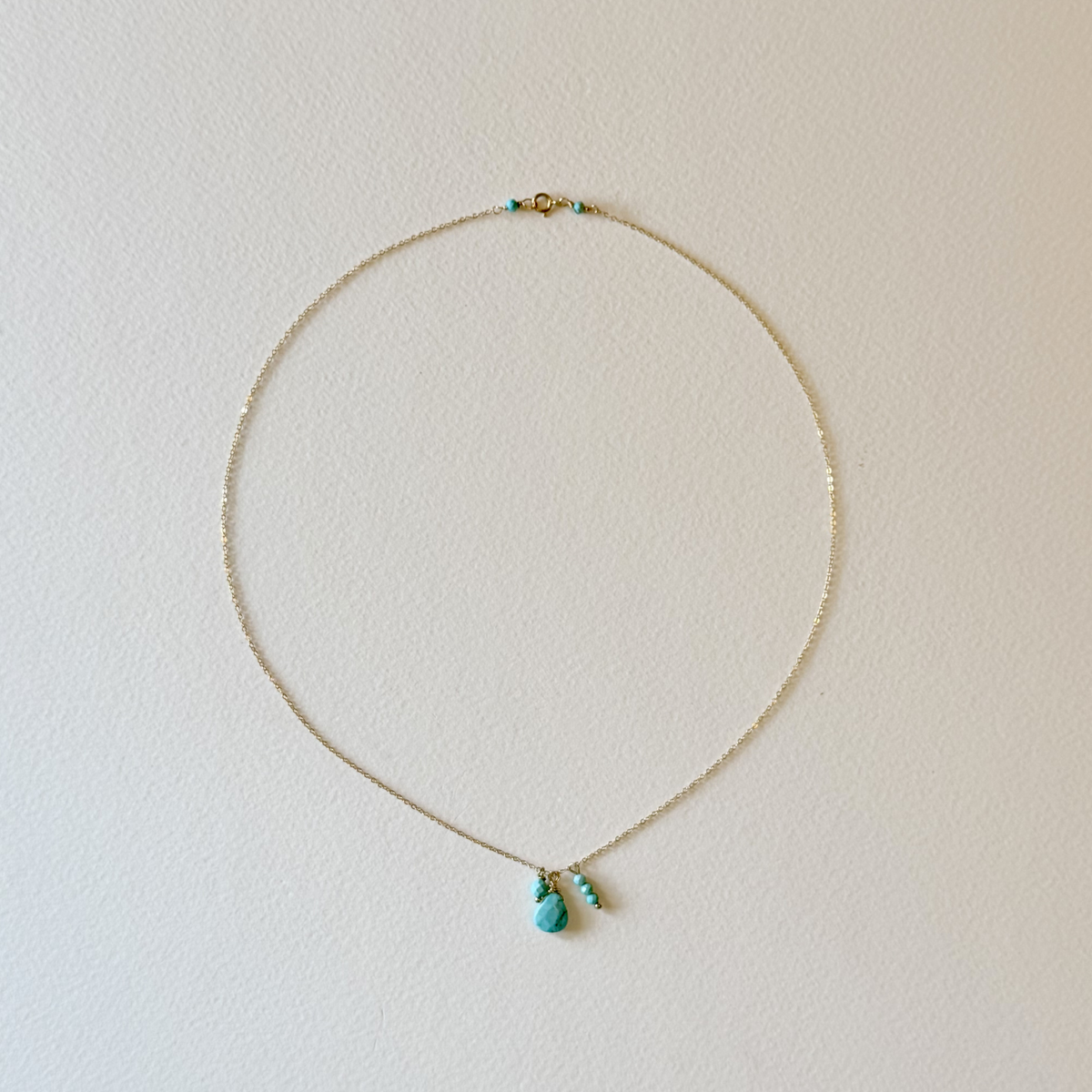 A delicate gold chain necklace with a small turquoise pendant and matching turquoise bead accents, displayed on a textured off-white background.
