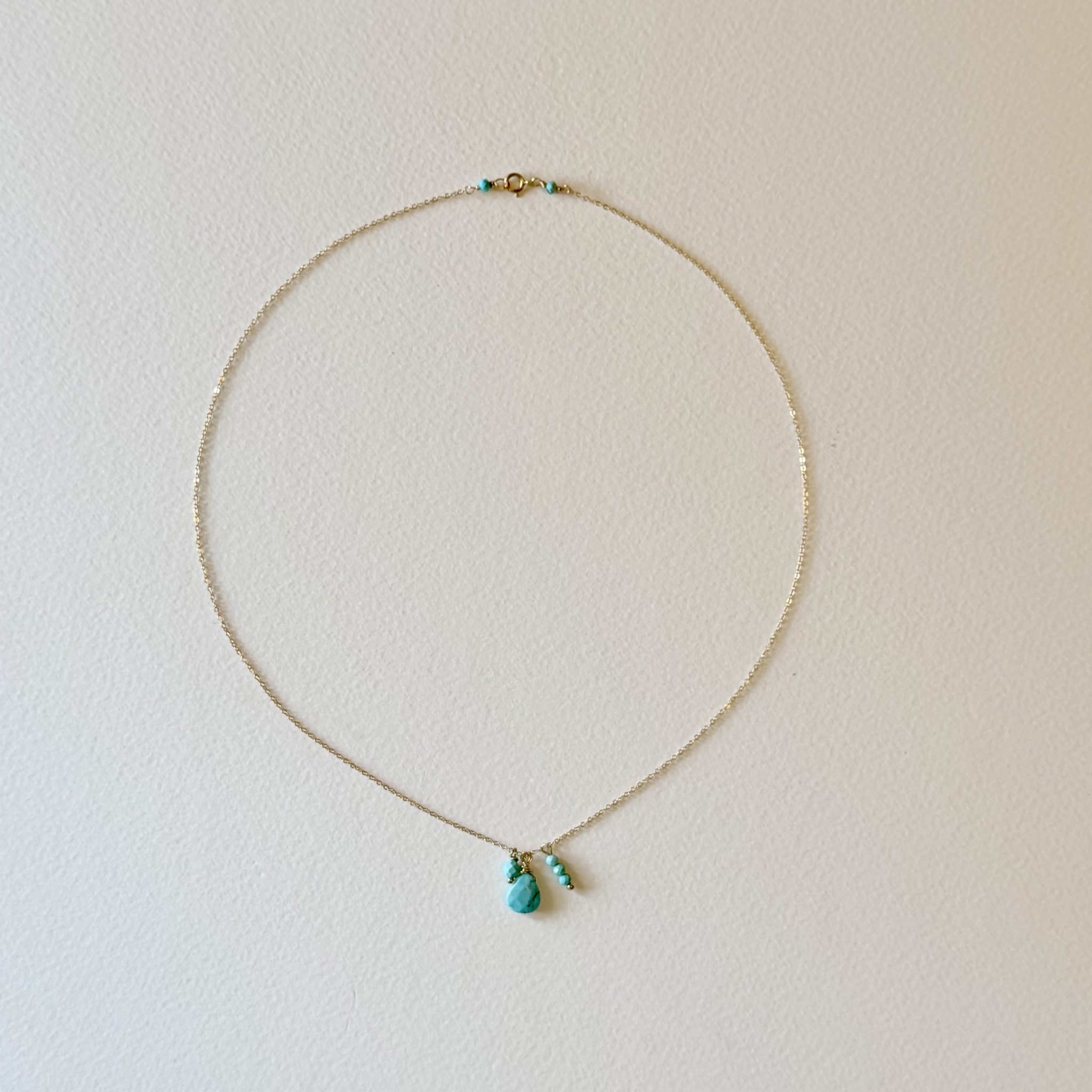 A delicate gold chain necklace with a small turquoise bead and a larger teardrop-shaped turquoise pendant, displayed on a textured off-white background.