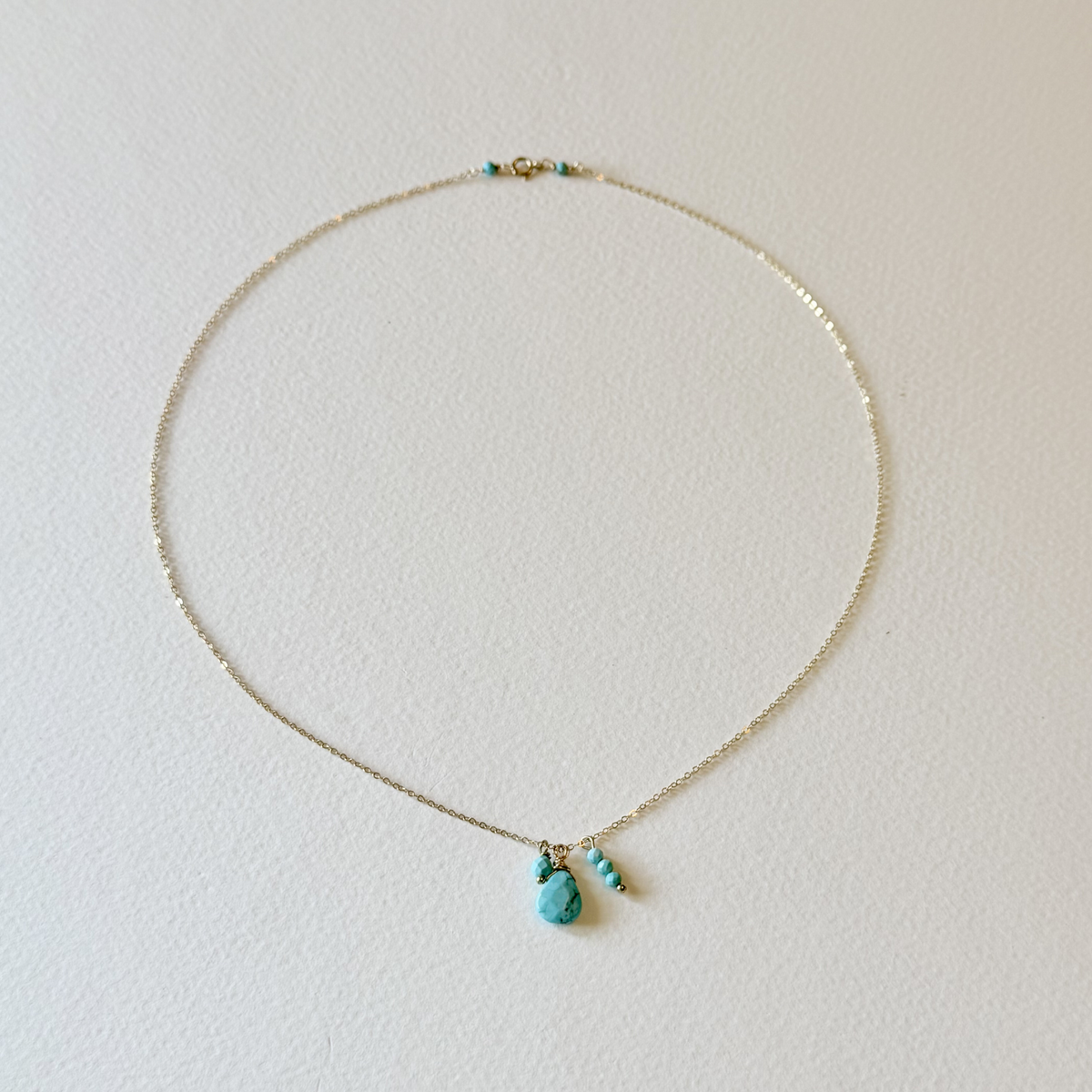 A delicate gold chain necklace with a small turquoise pendant and two tiny turquoise beads, displayed on a textured off-white background.