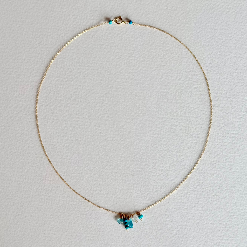 A delicate gold chain necklace with a small cluster of turquoise blue beads and a tiny gold charm at the center, displayed on a textured white background.