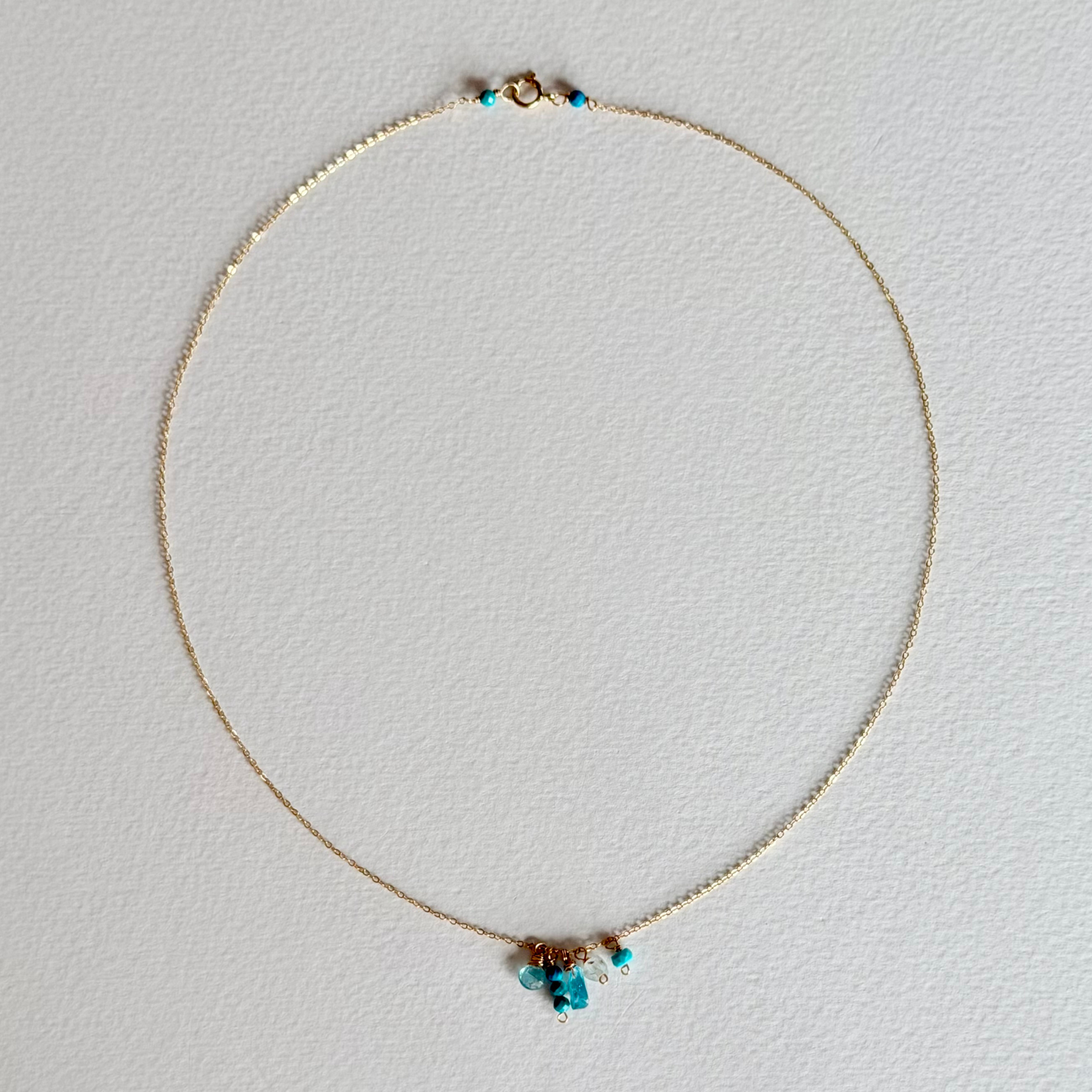 A delicate gold chain necklace with a small cluster of turquoise blue beads and a tiny gold charm at the center, displayed on a textured white background.