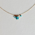 A delicate gold chain necklace featuring a small cluster of turquoise blue beads and a tiny heart charm, displayed on a textured white background.