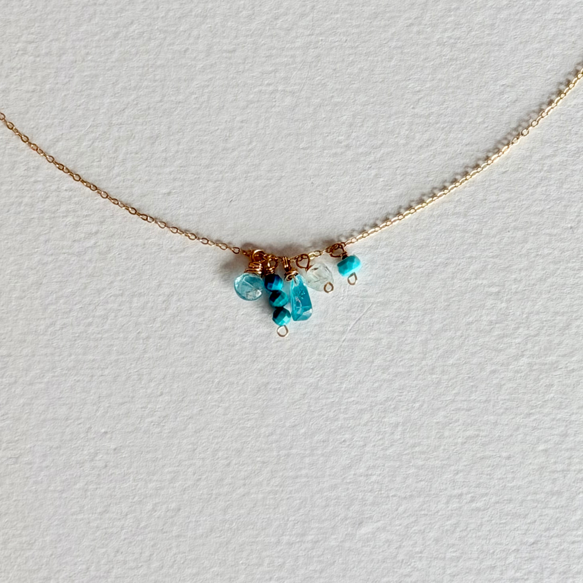 A delicate gold chain necklace featuring a small cluster of turquoise blue beads and a tiny heart charm, displayed on a textured white background.