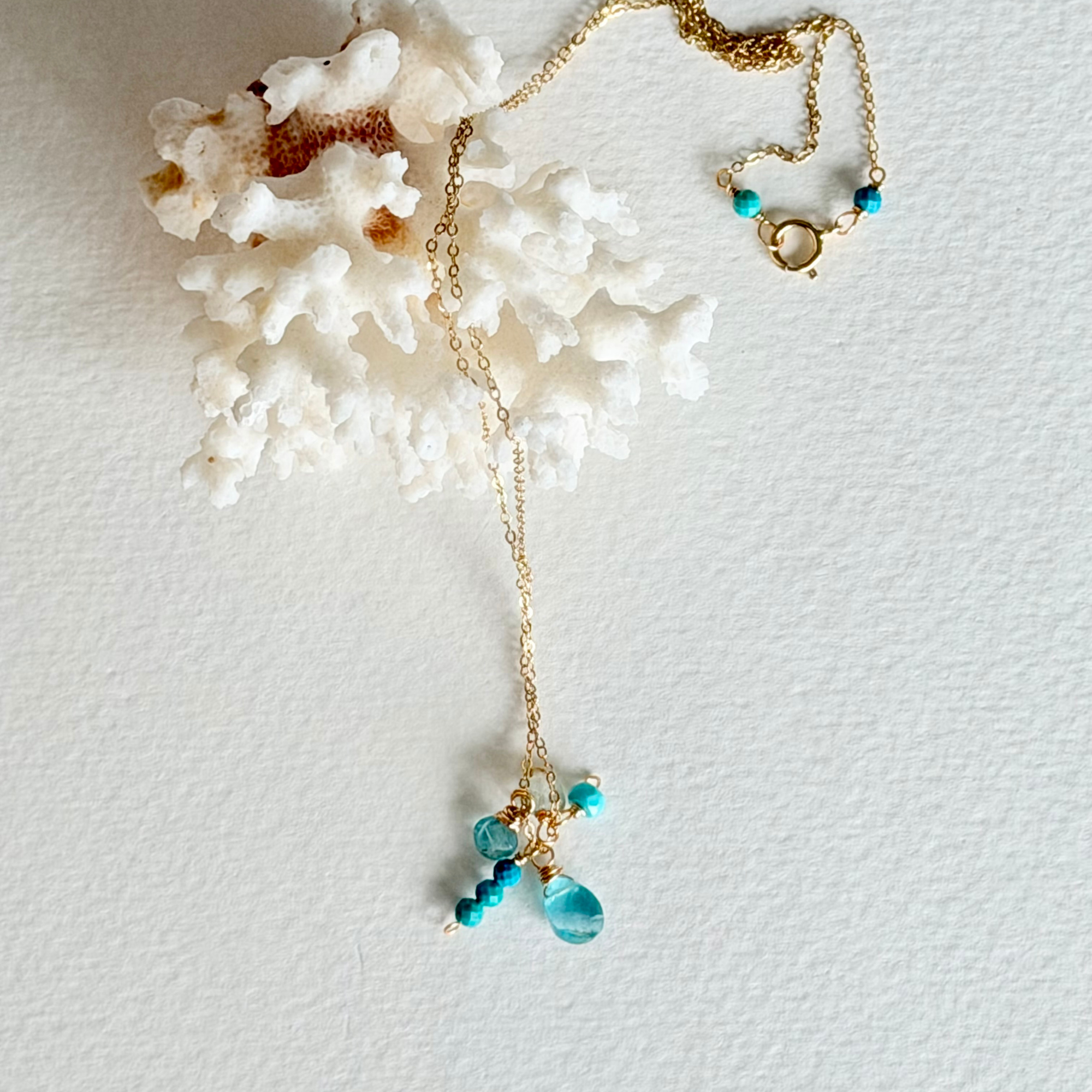 A delicate gold chain necklace with turquoise blue beads and a teardrop pendant, styled on a textured white background next to a white coral shell.