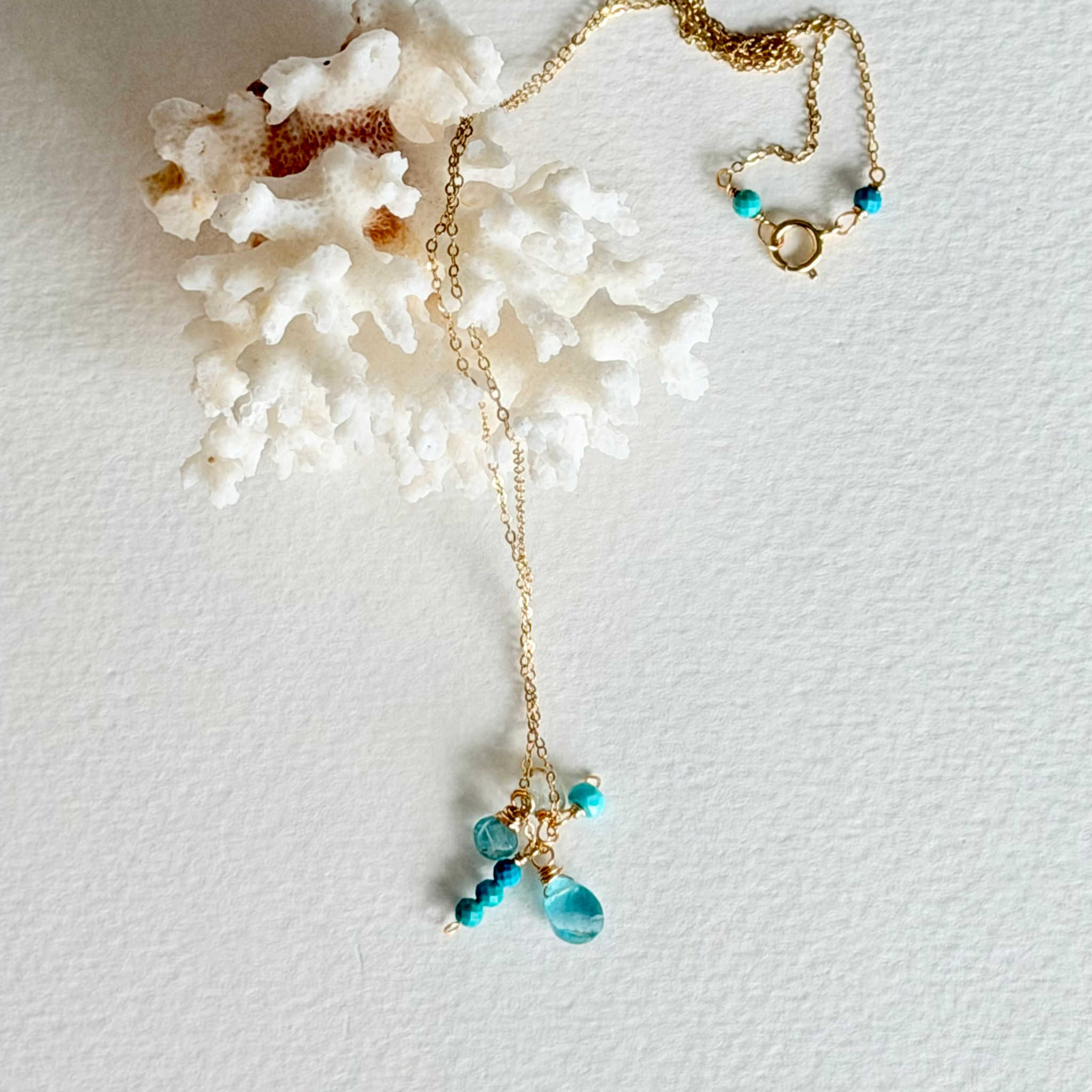 A delicate gold chain necklace with turquoise blue beads and a teardrop pendant, styled on a textured white background next to a white coral shell.
