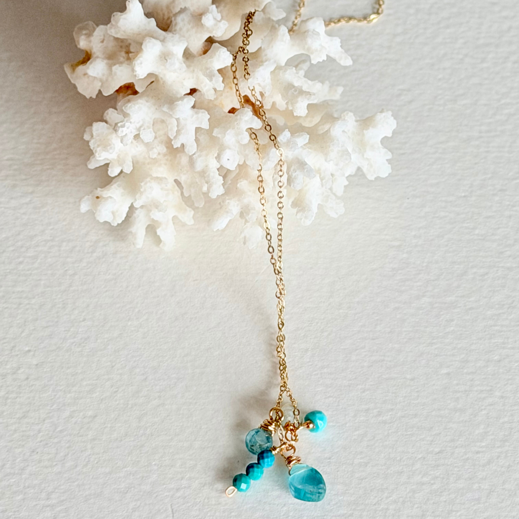 A delicate gold chain necklace with turquoise blue beads and a faceted blue teardrop pendant, displayed on a textured white background with a white coral prop.