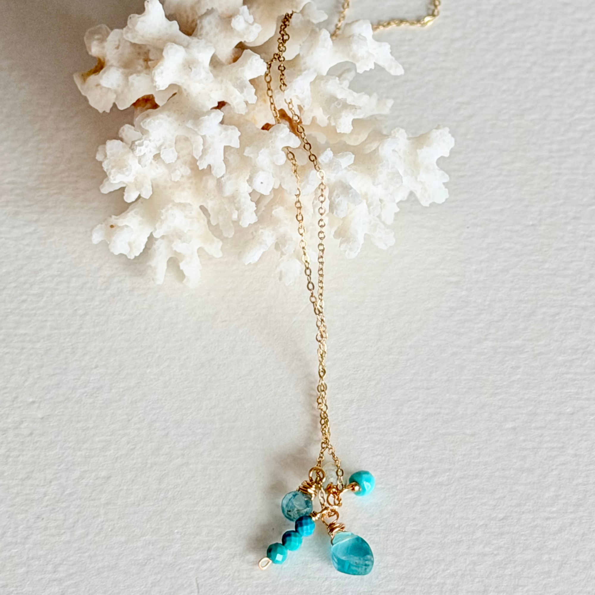 A delicate gold chain necklace with turquoise blue beads and a faceted blue teardrop pendant, displayed on a textured white background with a white coral prop.