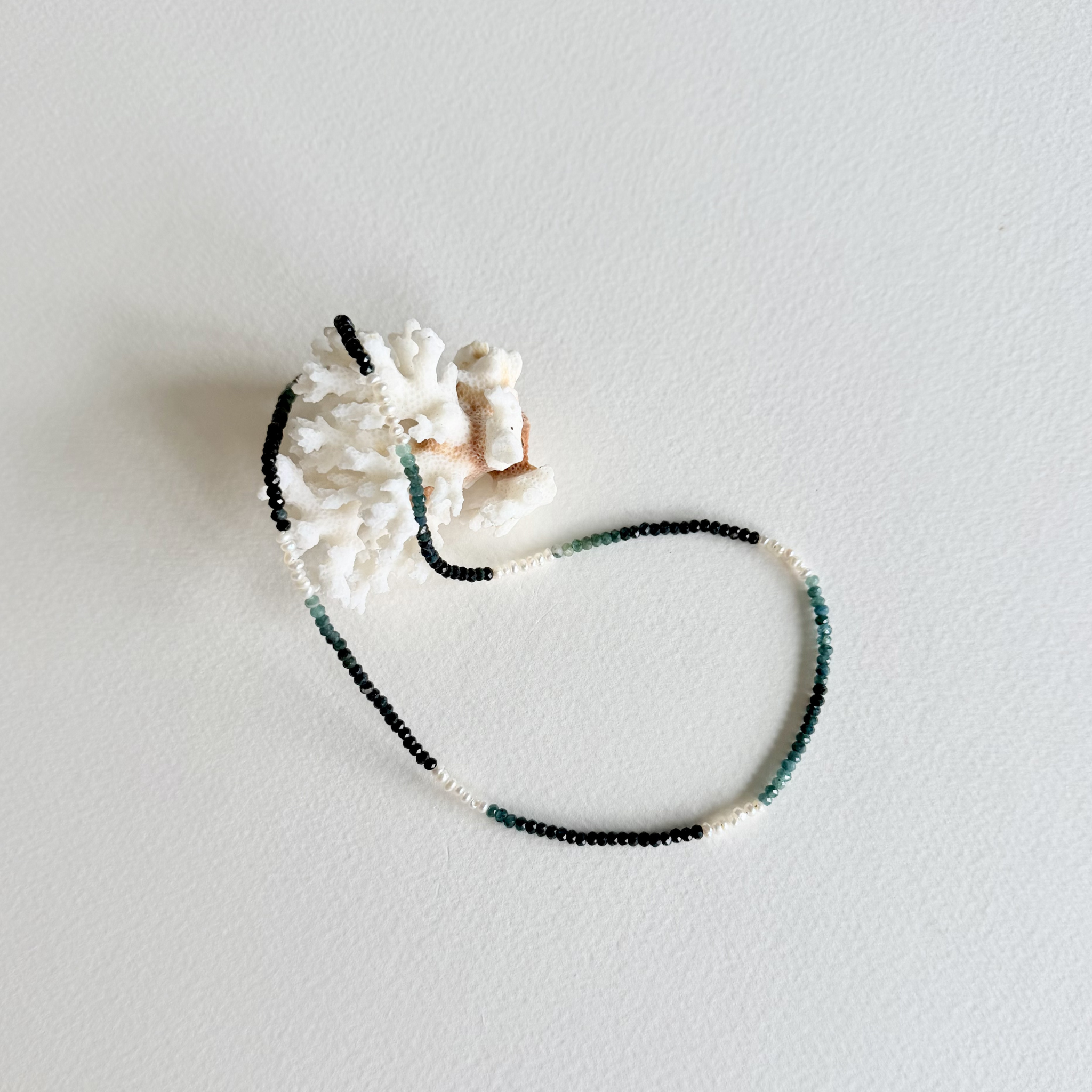A beaded necklace with black, white, and green beads, displayed on a white textured surface next to a white seashell.