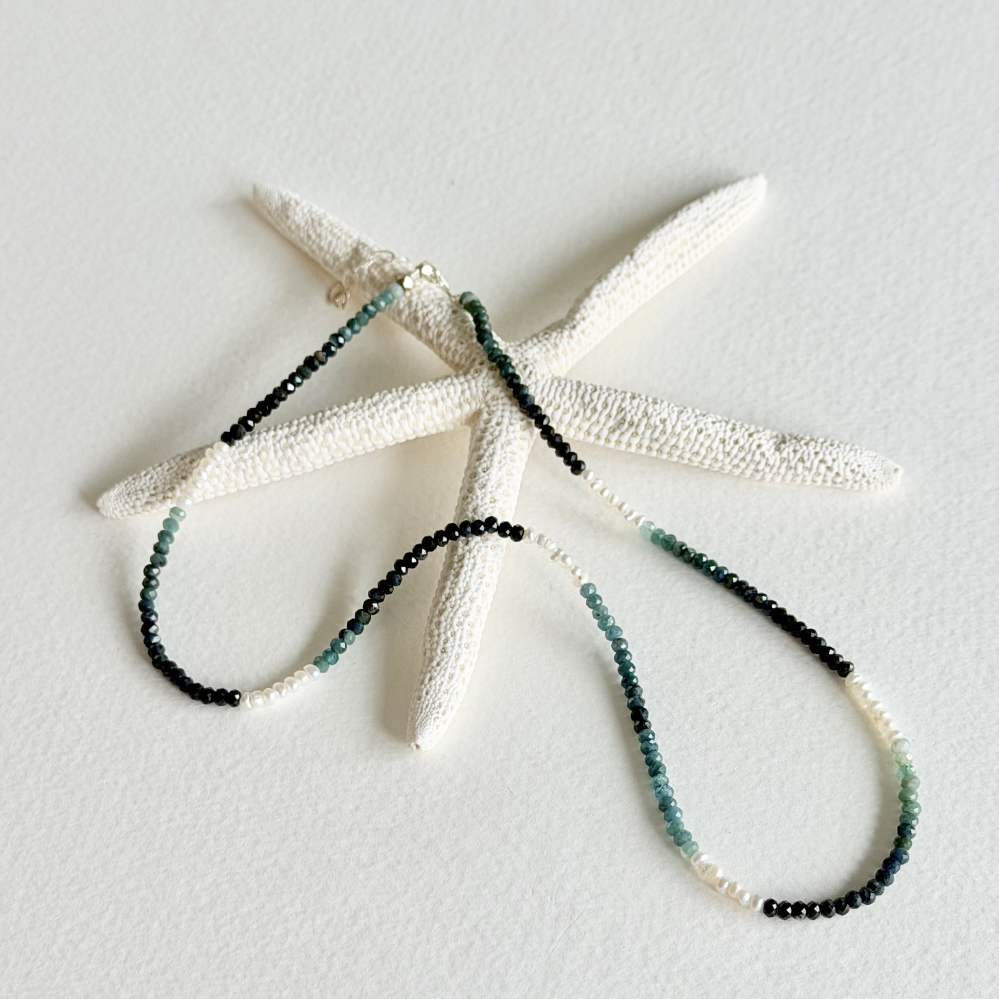 A beaded necklace with black, teal, and white beads draped over a white starfish prop on a textured white background.
