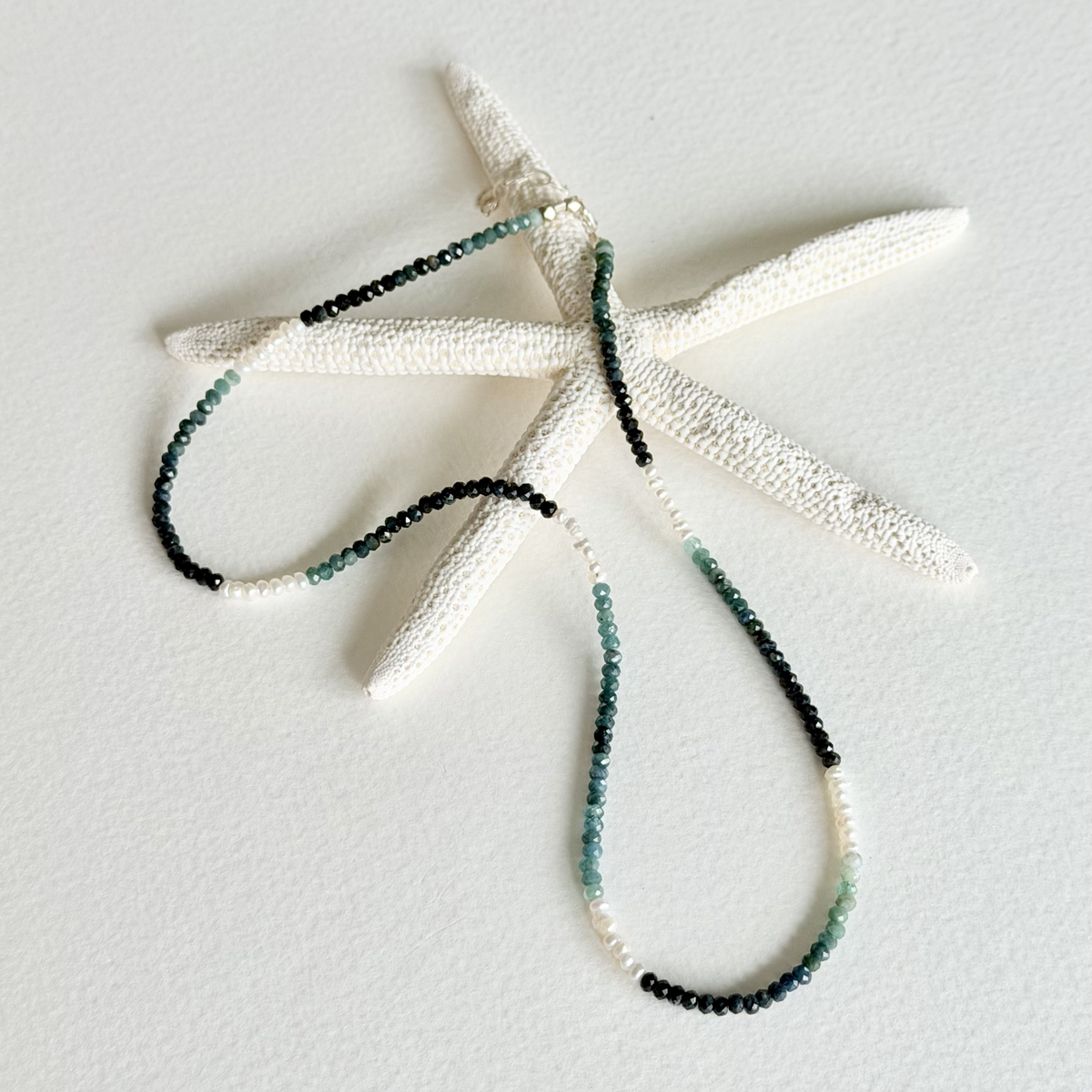 A beaded necklace with alternating black and green beads draped over a white starfish prop on a textured white background.