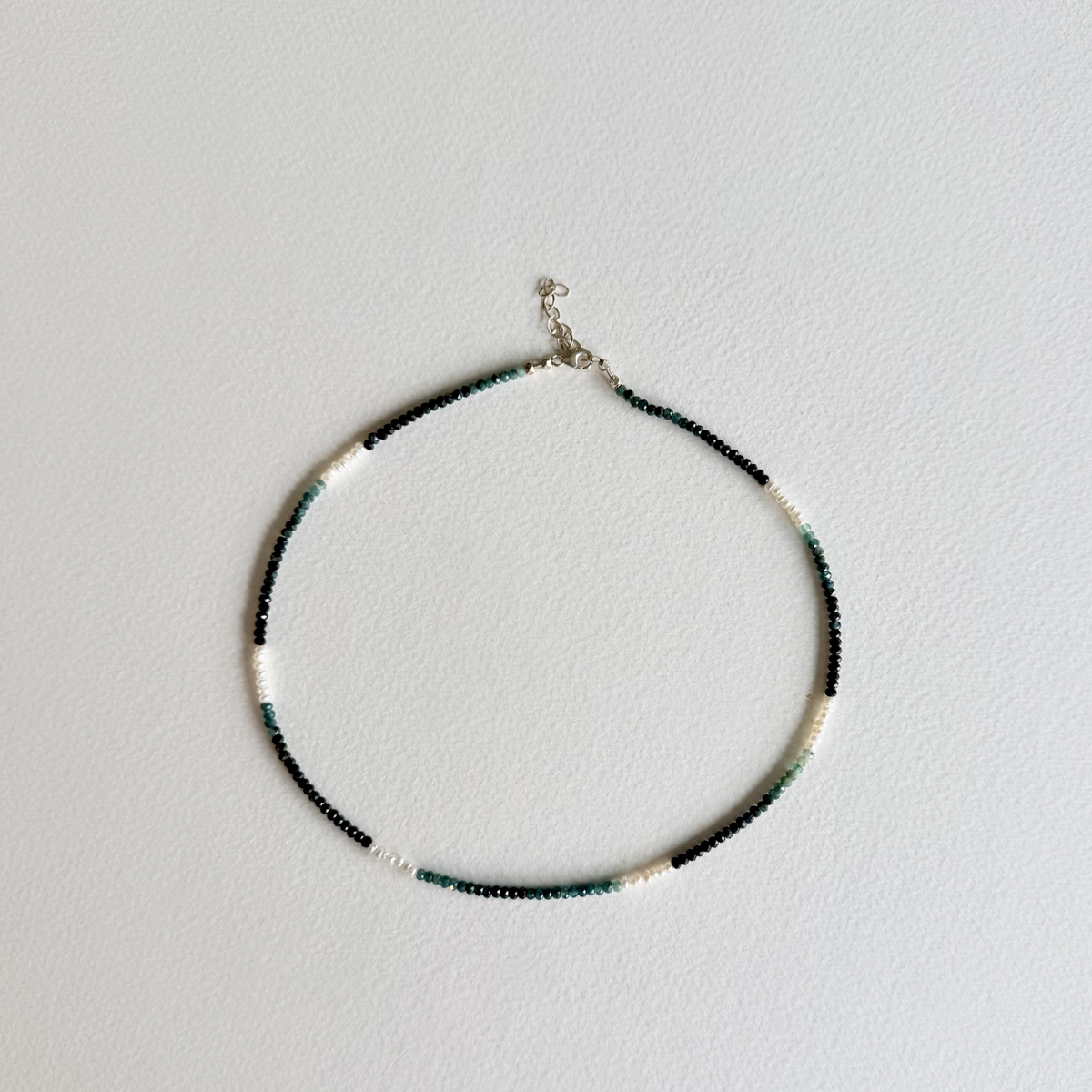 A delicate beaded necklace with alternating sections of black, white, and dark green beads, featuring a silver-tone clasp, displayed on a light gray textured background.