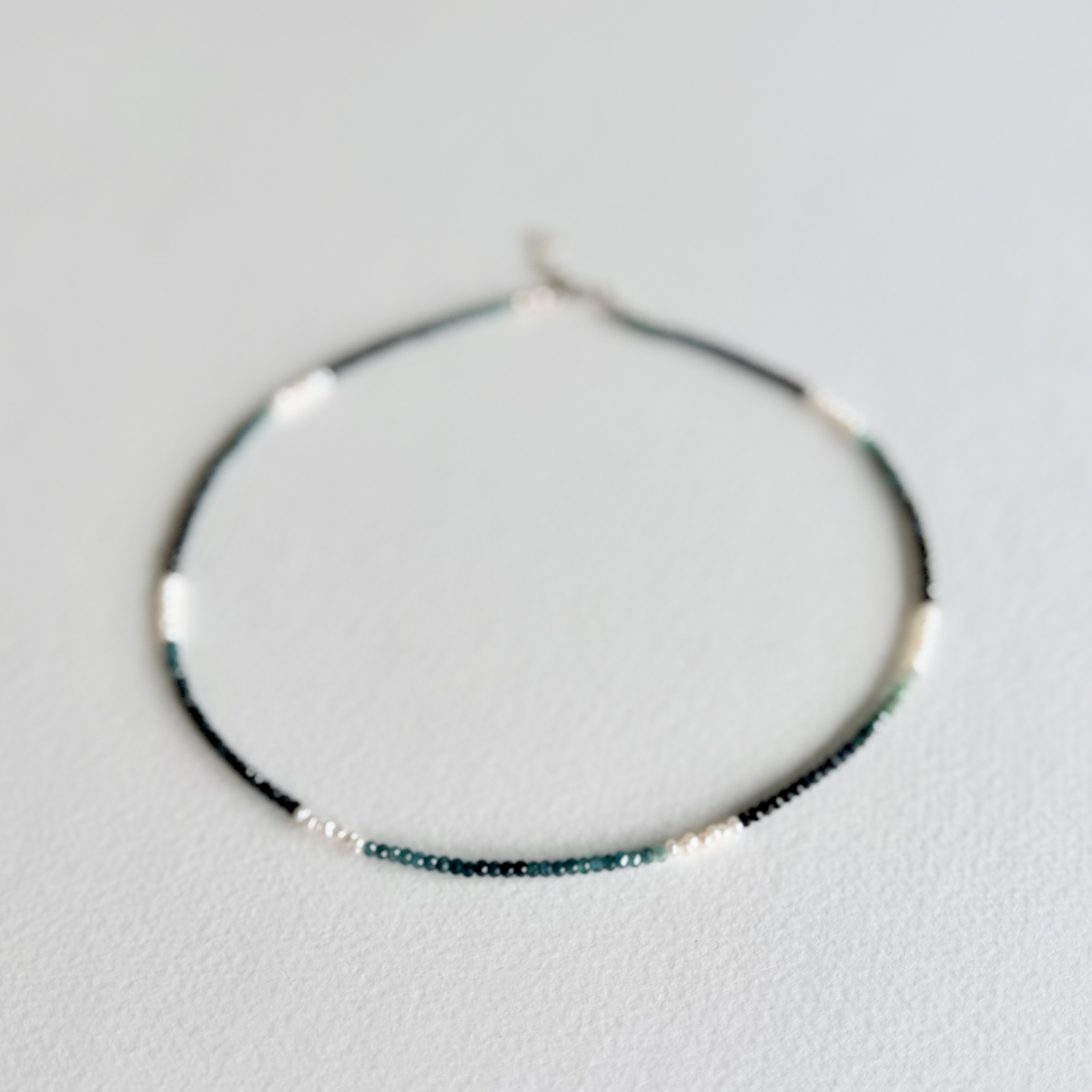 A delicate beaded necklace with small dark green and white beads, photographed on a light gray textured background.