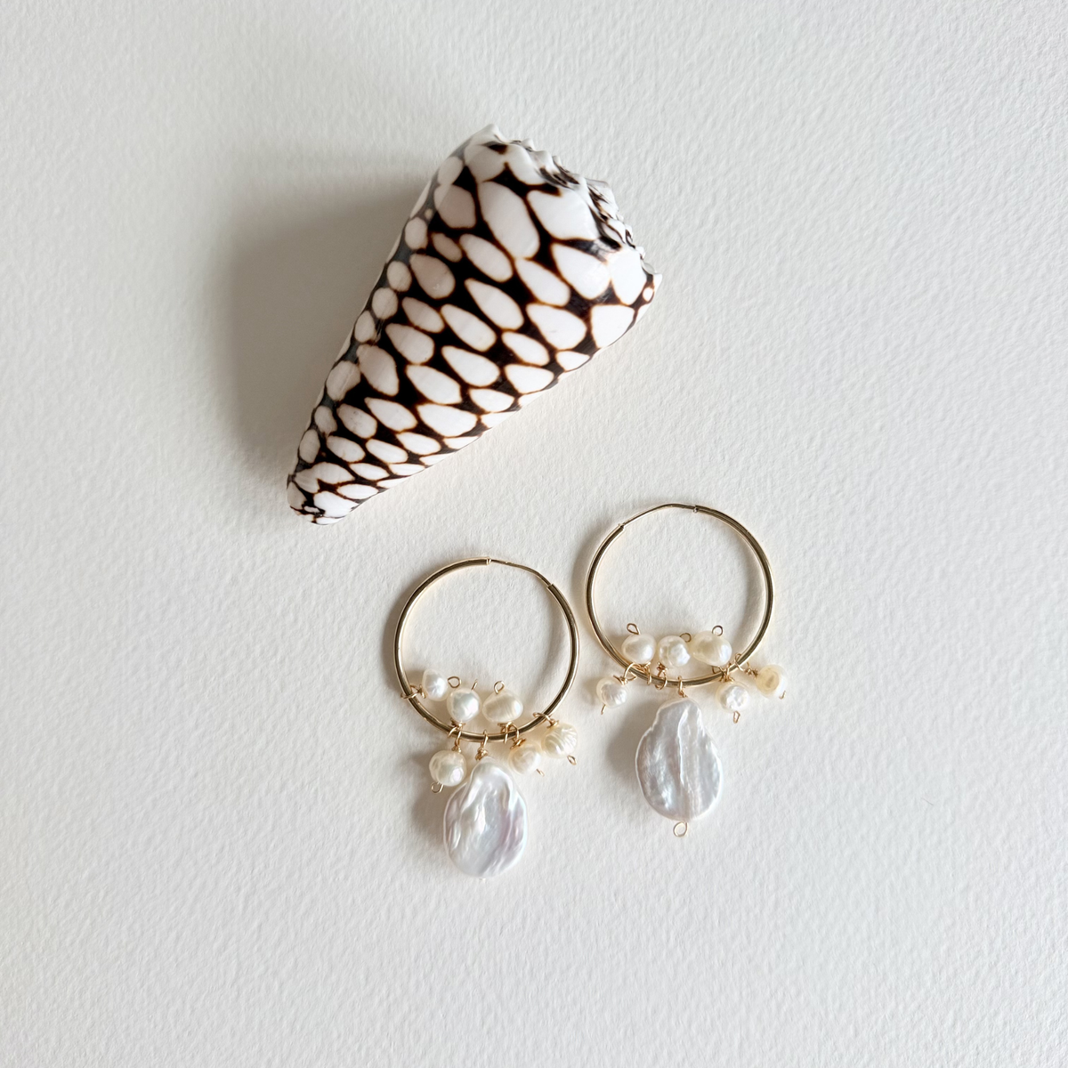 Pair of gold hoop earrings with pearl drops next to a shell on a light gray background