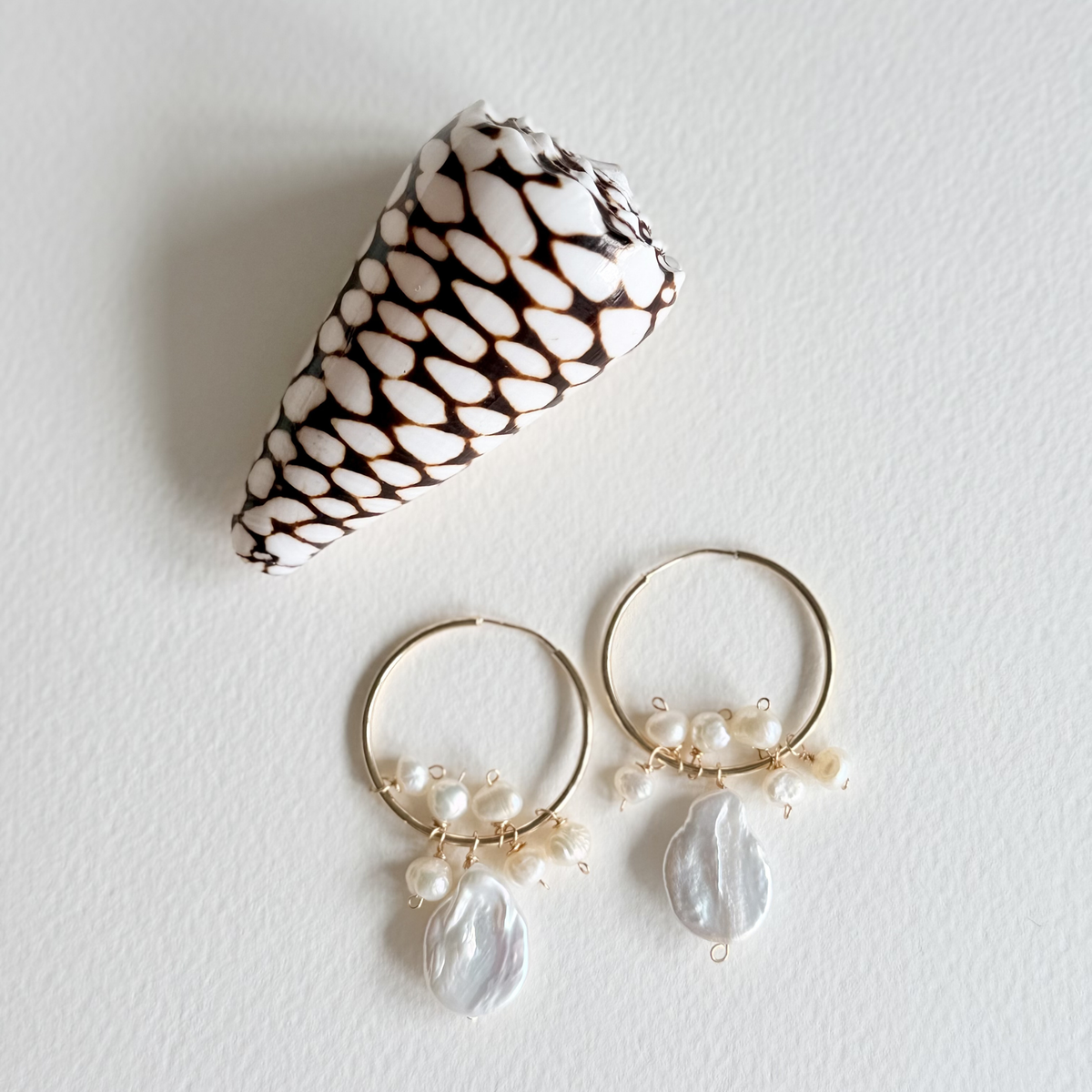 Pair of hoop earrings with pearl drops next to a shell on a light background