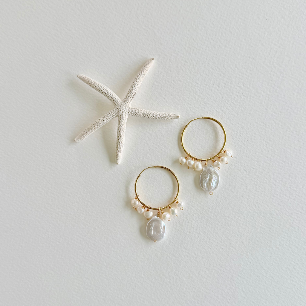 Gold hoop earrings with pearls and shell charms on a light gray background with a starfish.