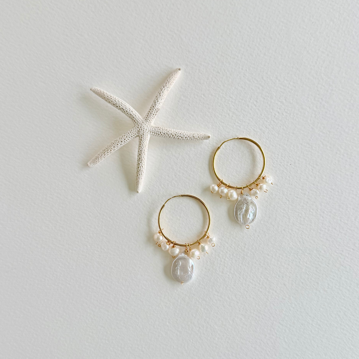 Gold hoop earrings with pearls and shell charms on a light gray background with a starfish.