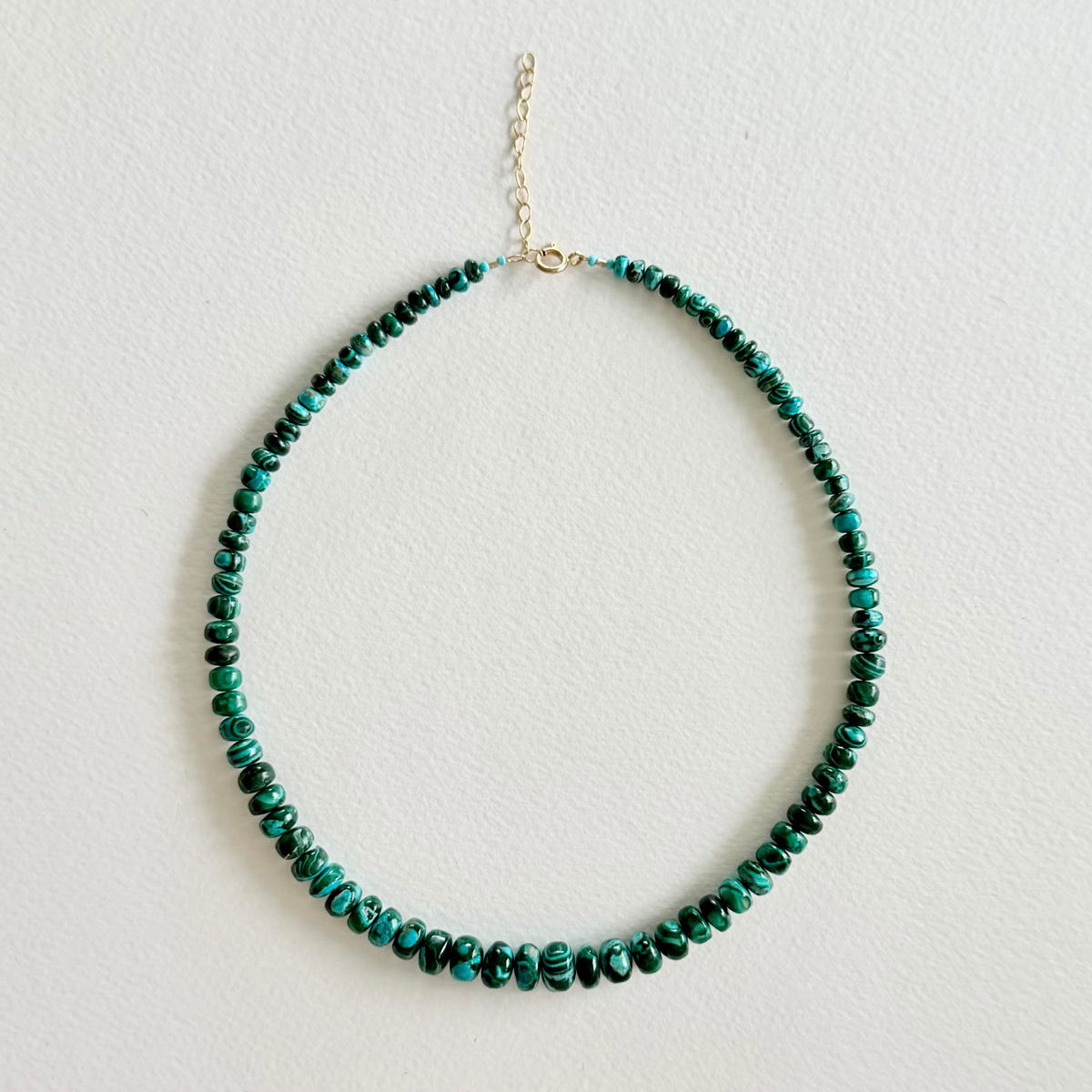 A beaded necklace made of small round green and turquoise malachite beads with a gold-tone clasp and extender chain, displayed against a light off-white textured background.