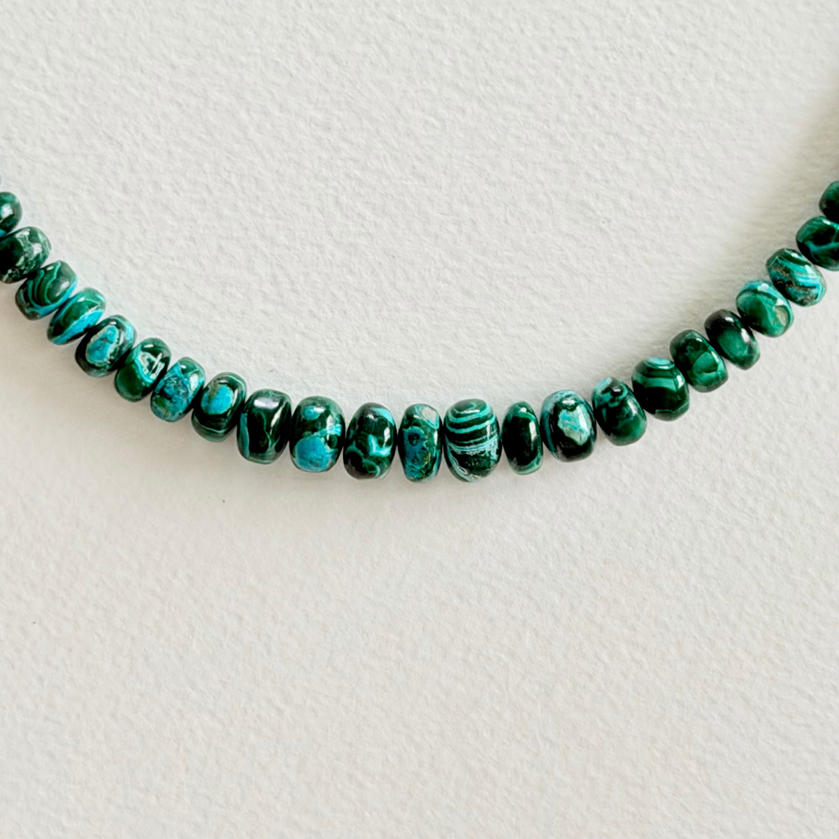 A close-up of a strand of polished green malachite beads with natural black and turquoise veining, arranged in a gentle curve on a textured off-white background.