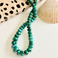 A beaded necklace made of polished green malachite beads, styled on a white surface with a seashell and a black-and-beige spotted fabric prop.