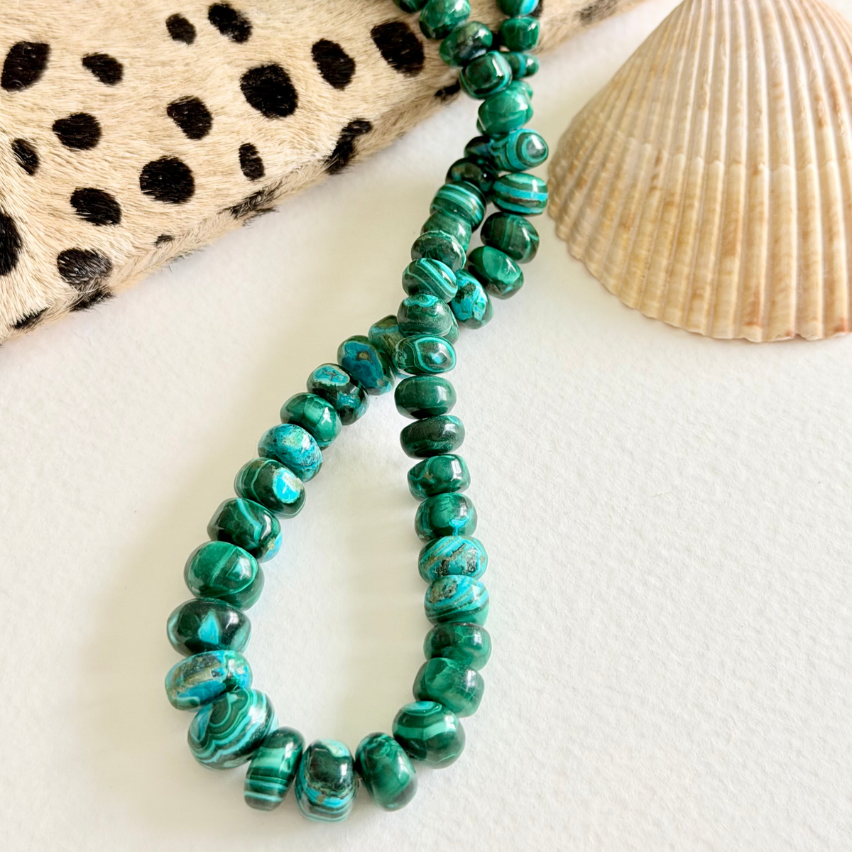 A beaded necklace made of polished green malachite beads, styled on a white surface with a seashell and a black-and-beige spotted fabric prop.