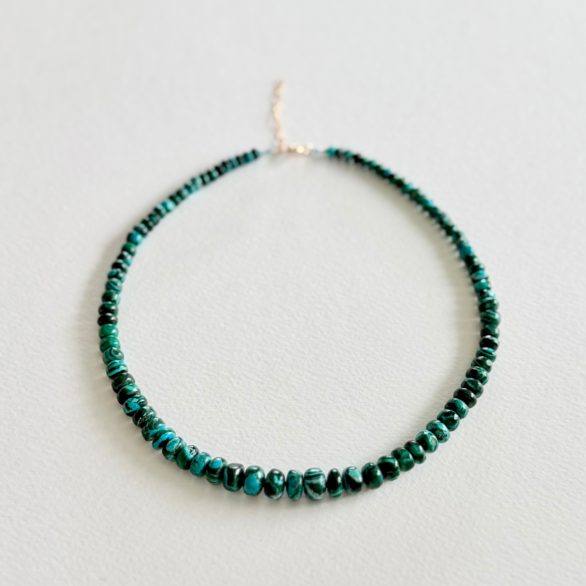 A beaded necklace made of small dark green and turquoise beads with a gold-tone clasp, displayed on a light gray textured background.