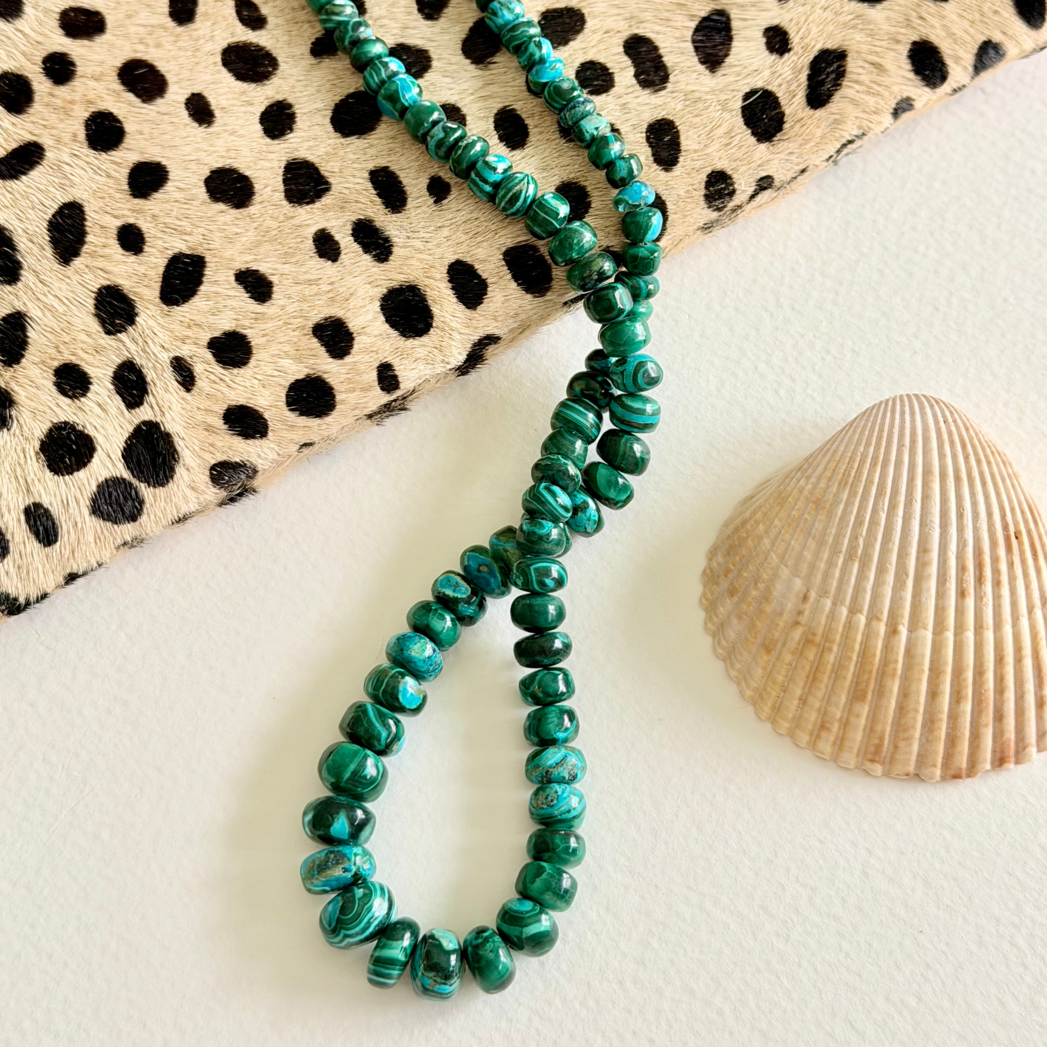 A beaded necklace made of glossy green malachite  round beads draped on a white surface, styled with a leopard-print fabric and a seashell.