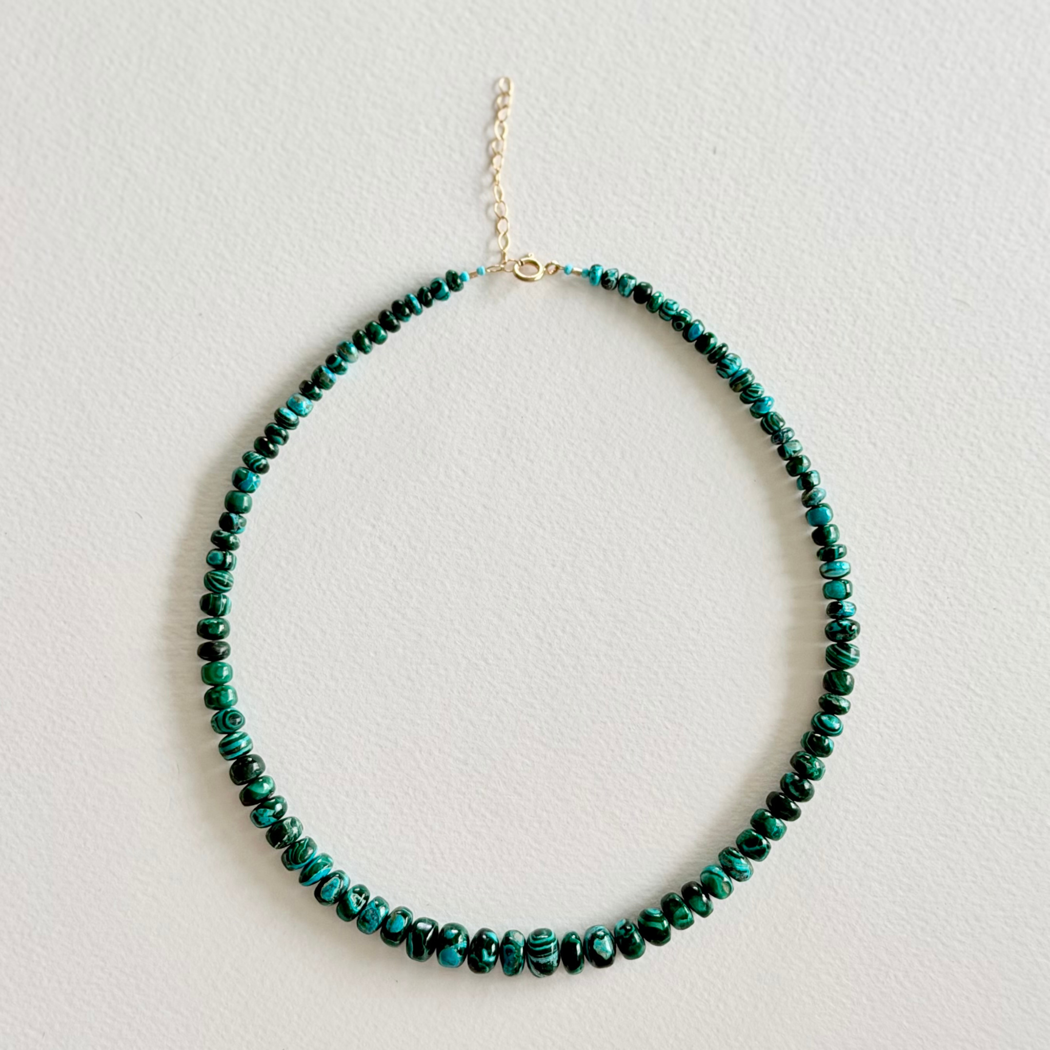 A beaded necklace made of small round beads in green and black, with a gold-tone chain extender and clasp, displayed on a light textured background.