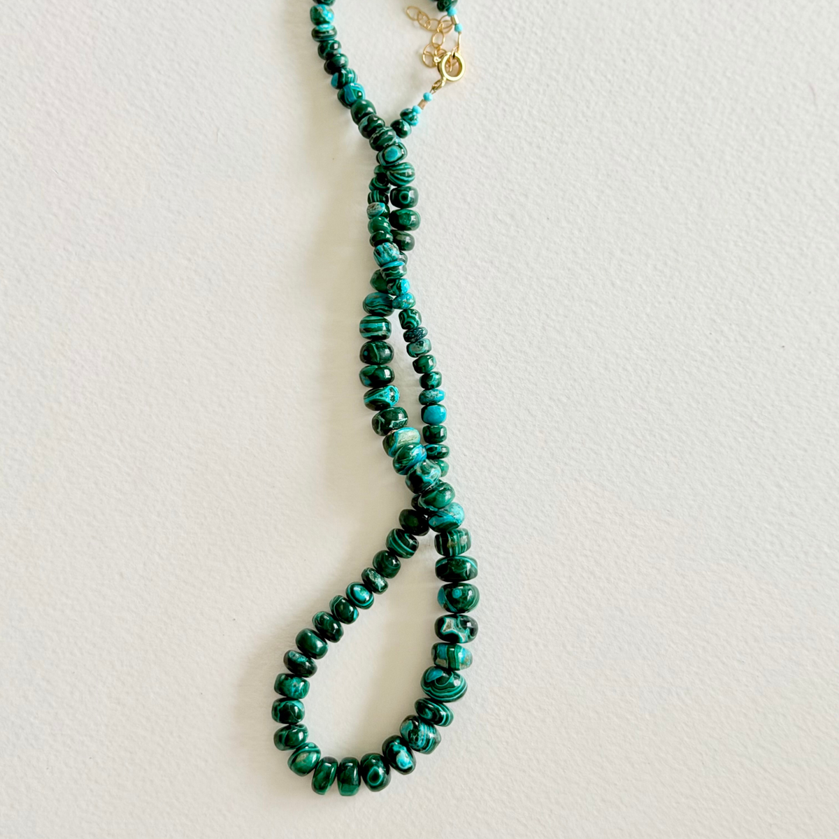 A beaded necklace made of dark green and turquoise marbled beads, arranged in a loop with a small gold clasp, photographed against a plain off-white background.