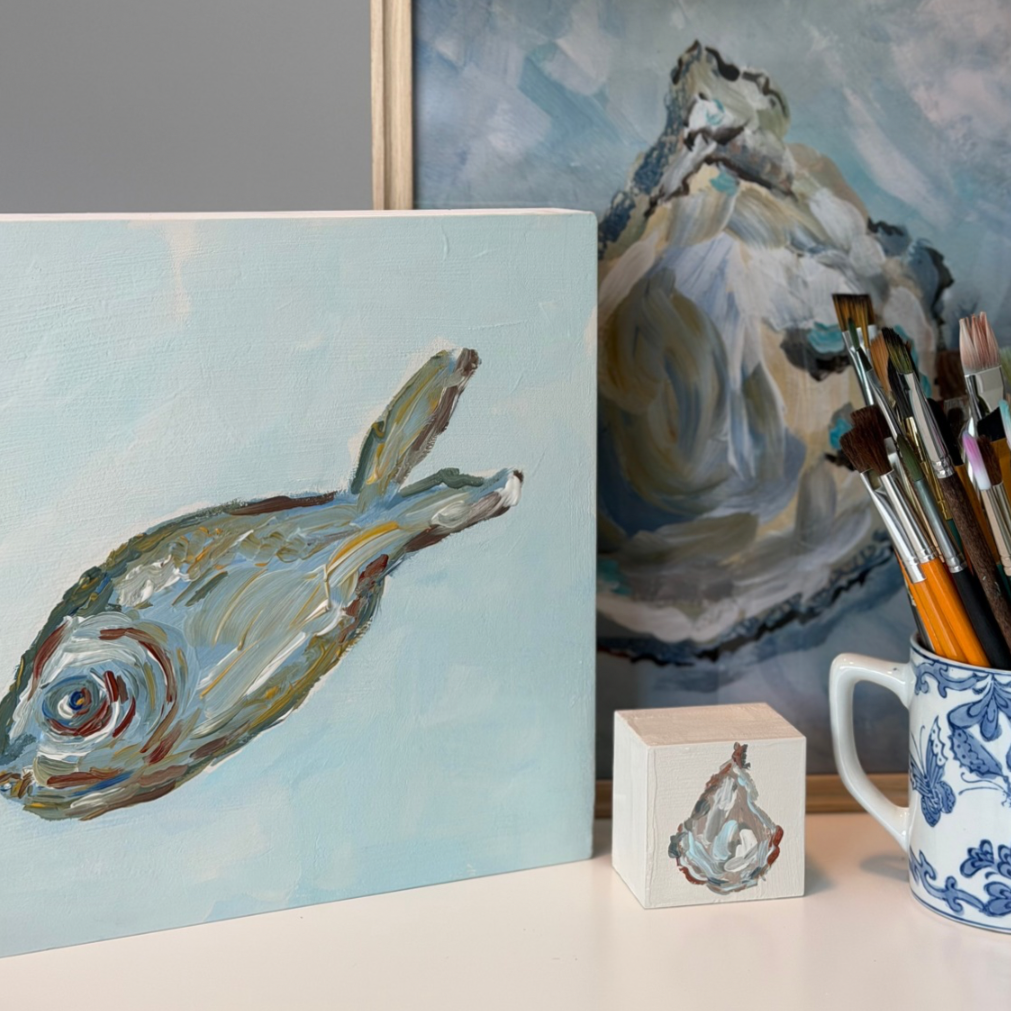 A painted canvas showing a fish on a pale blue background, displayed on a tabletop beside a mug filled with paintbrushes and another artwork in the background.