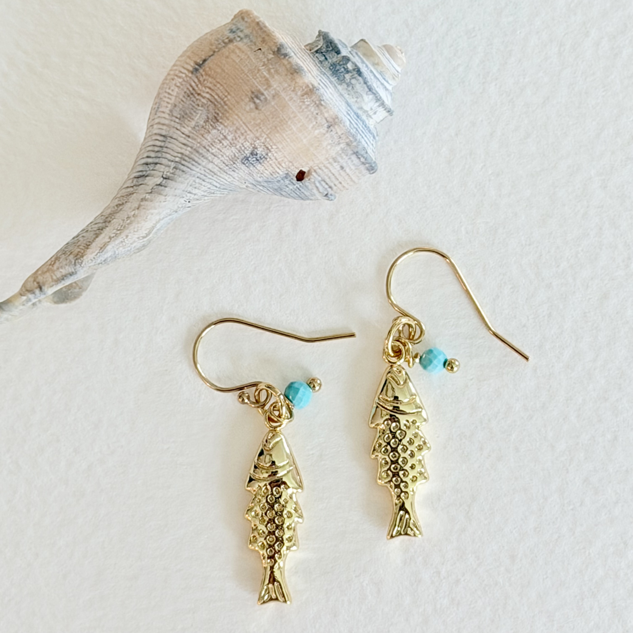Gold fish-shaped earrings with turquoise beads on a white background with a shell.
