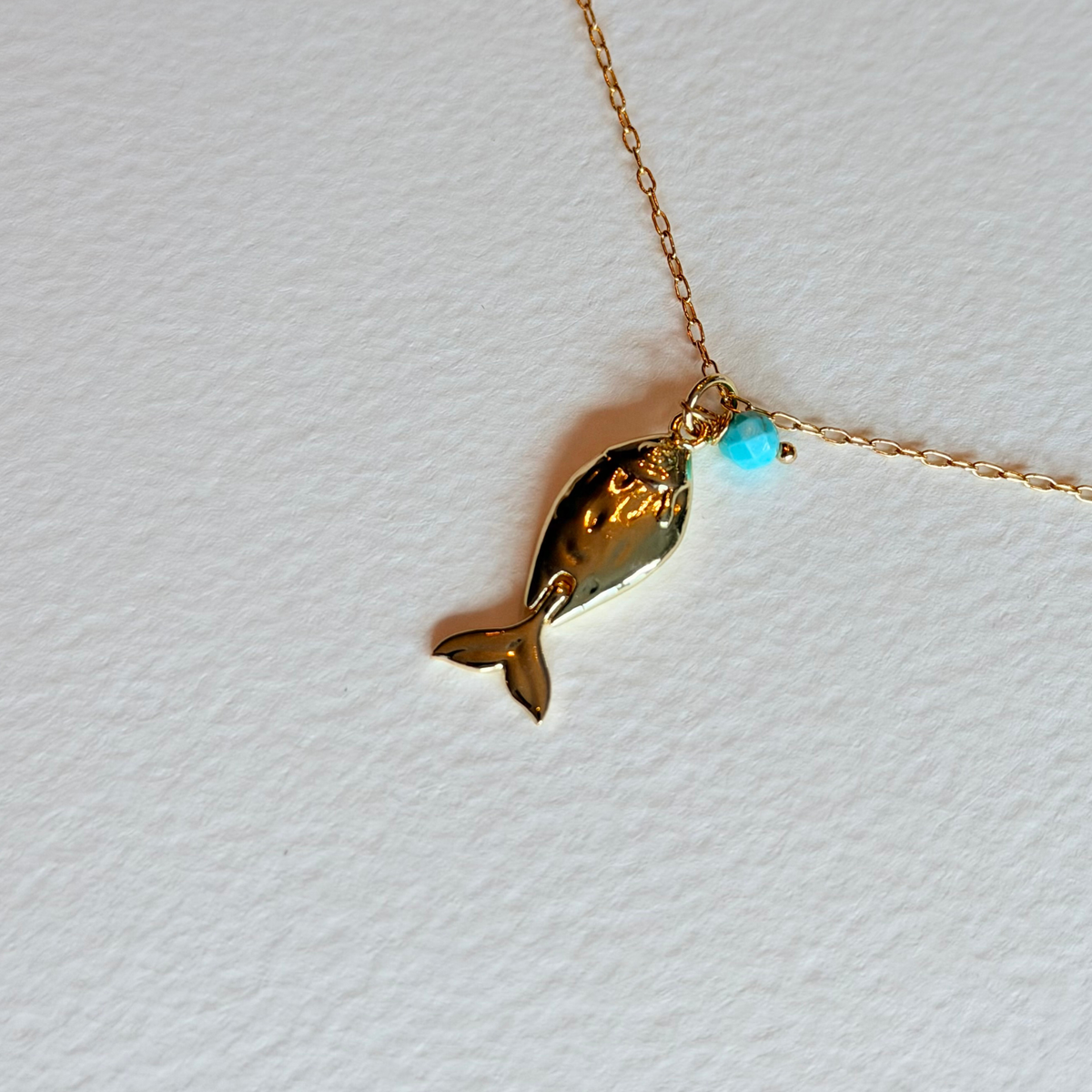 Gold fish-shaped pendant on a chain with a light blue gemstone against a white background