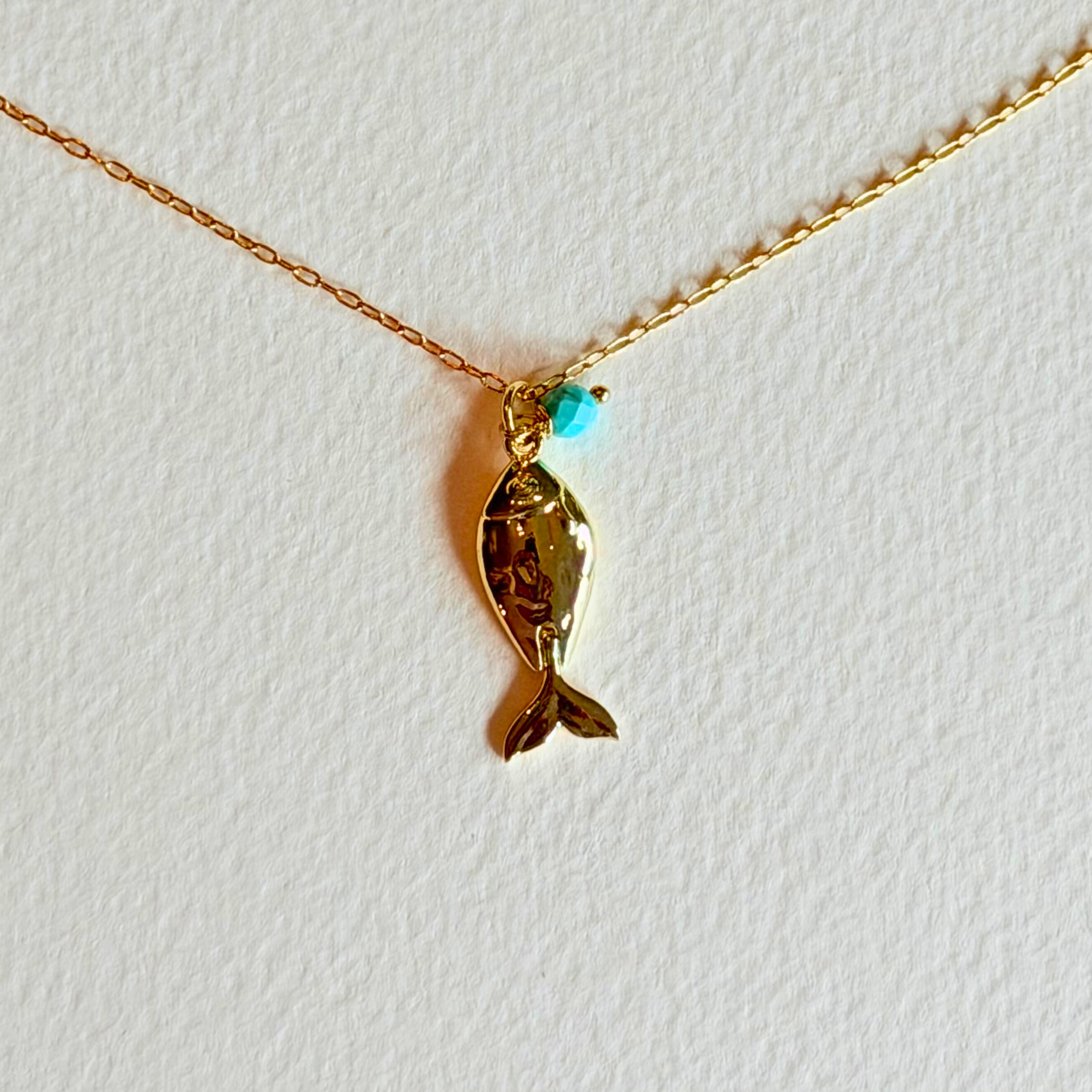 Gold fish-shaped pendant with a turquoise gemstone on a gold chain against a white background