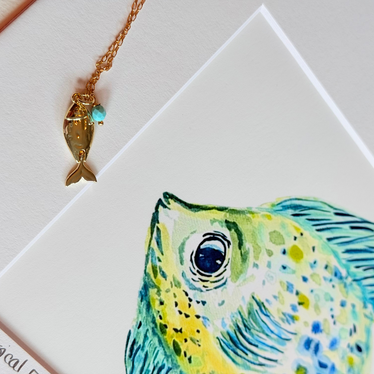 Gold necklace with a fish pendant on a card featuring a colorful fish illustration.