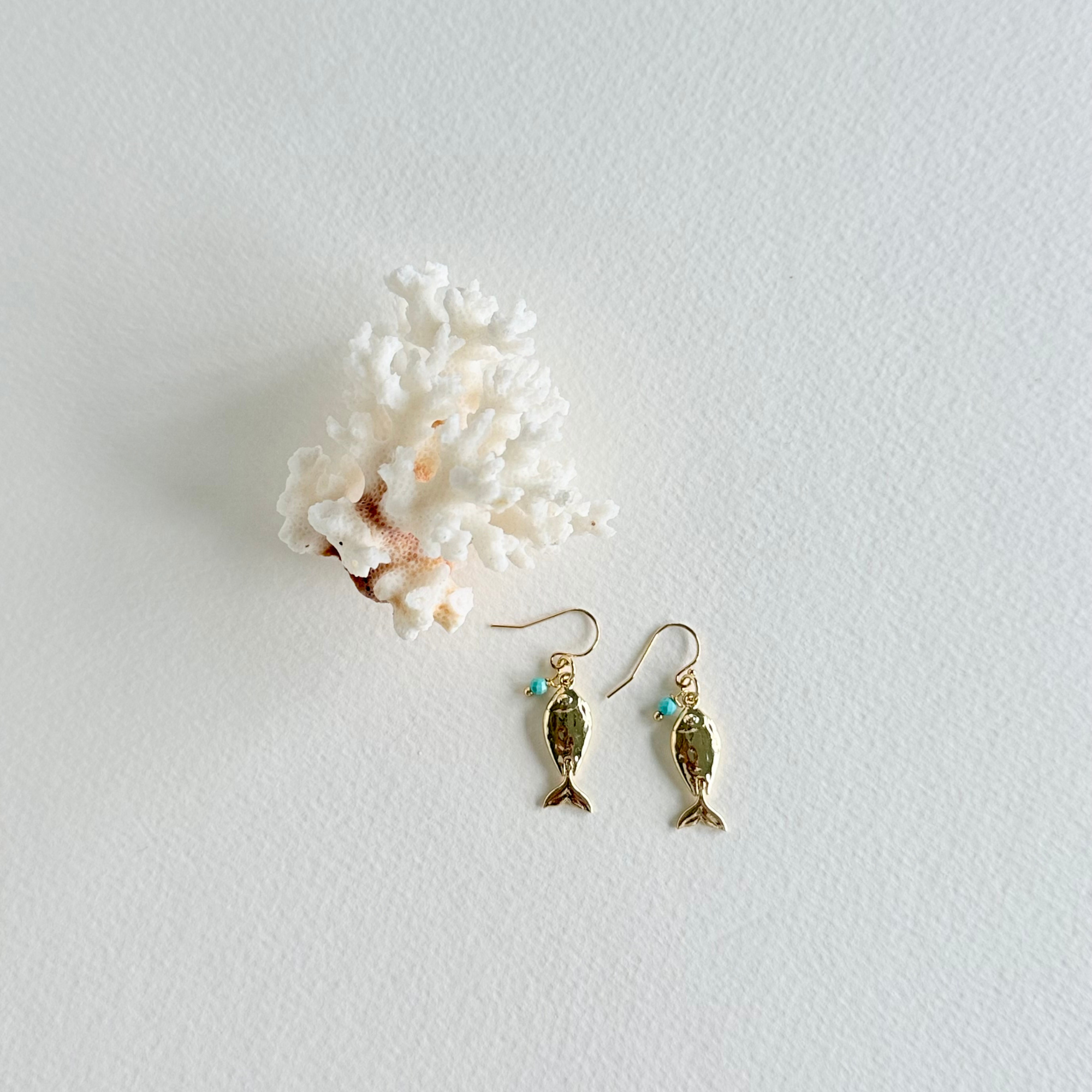 Gold fish-shaped earrings with turquoise stones on a light gray background with white coral.