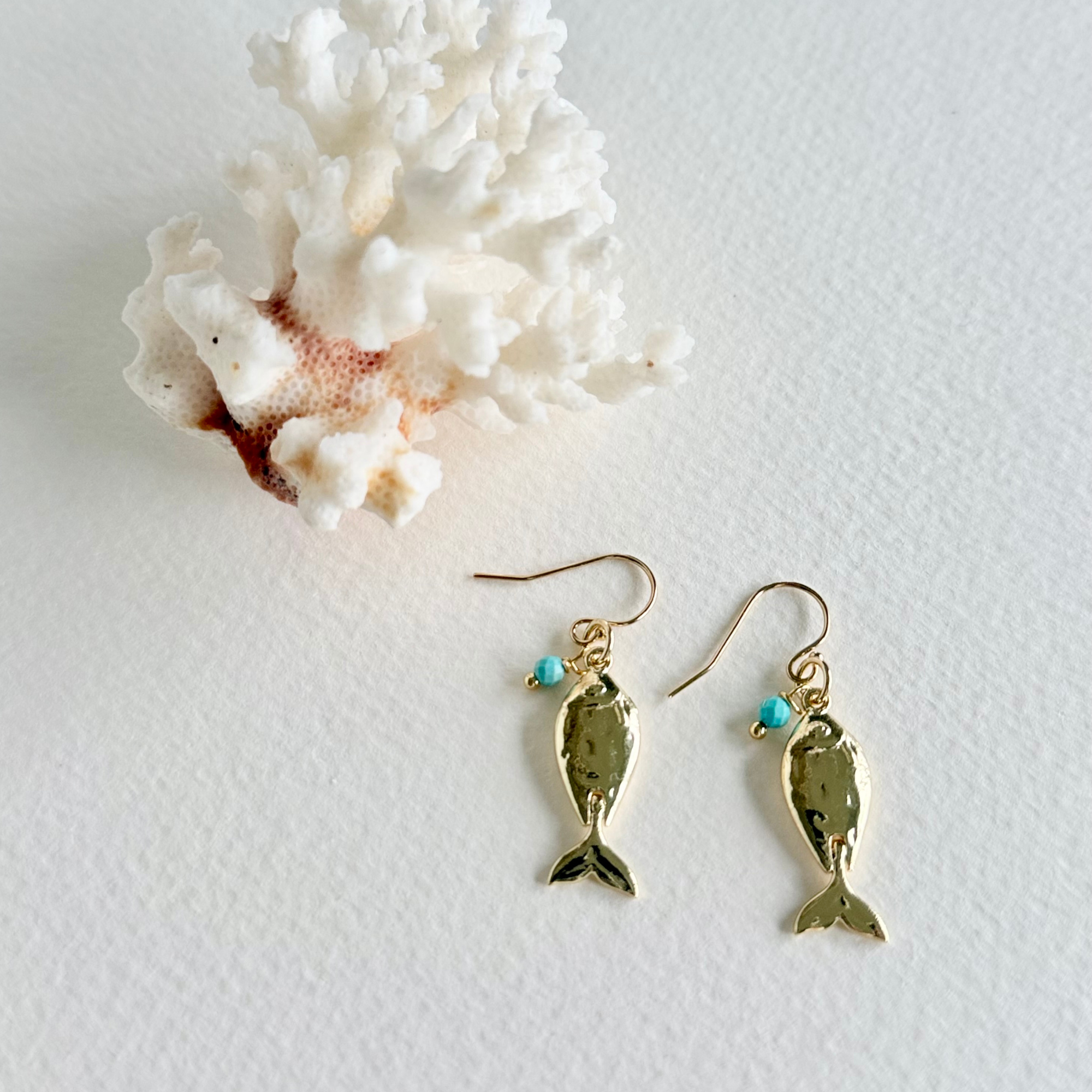 Gold fish-shaped earrings with turquoise beads on a white background with coral.