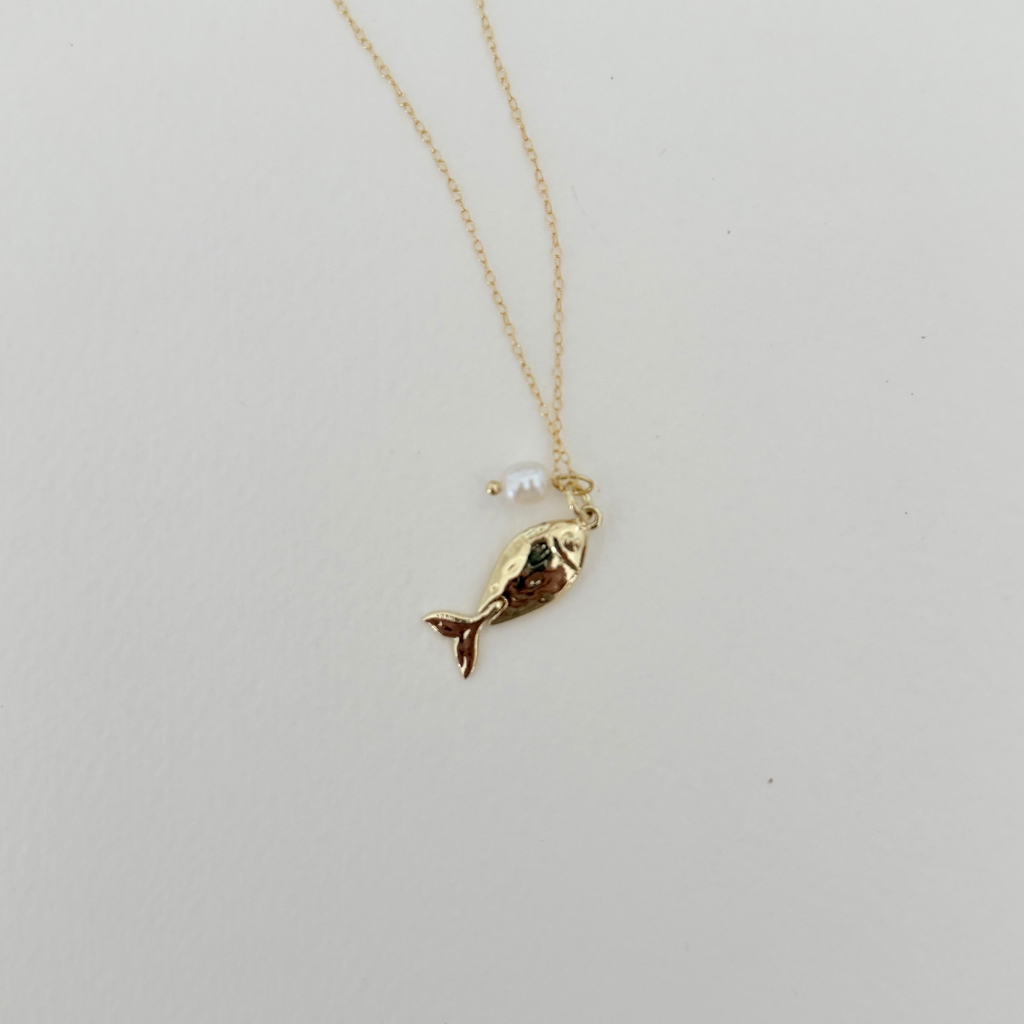 A delicate gold chain necklace featuring a small gold fish charm with a tiny white pearl bead hanging beside it, displayed on a clean white background.