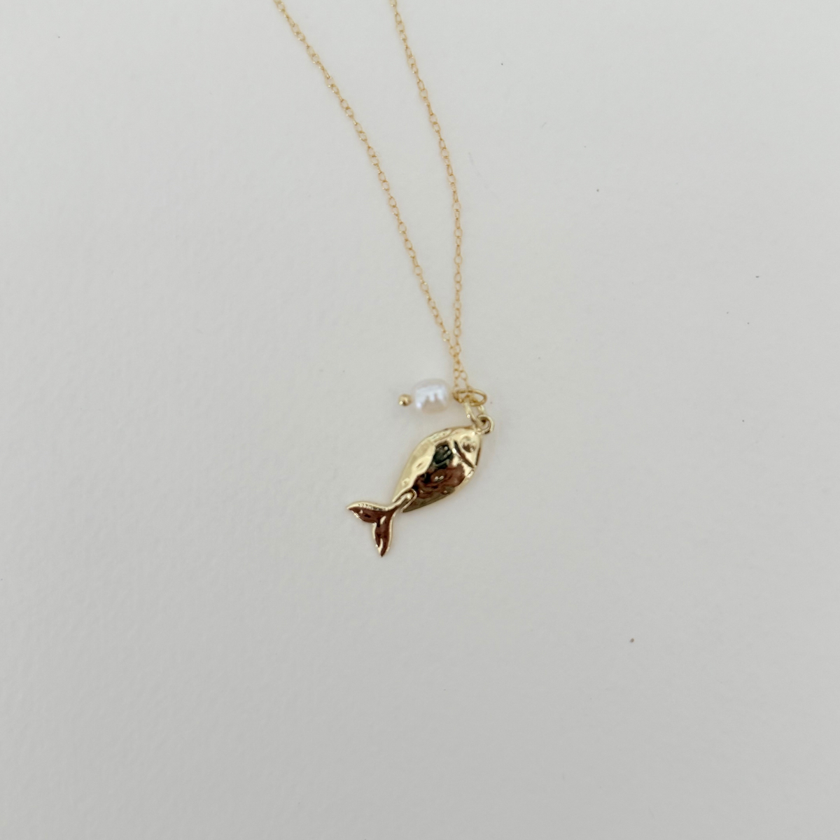 A delicate gold chain necklace featuring a small gold fish charm with a tiny white pearl bead hanging beside it, displayed on a clean white background.