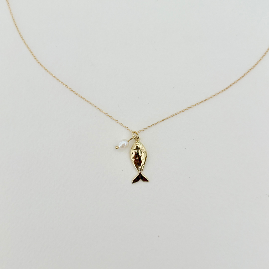 A delicate gold chain necklace featuring a small gold fish pendant with a dark tail and a tiny white pearl bead, displayed against a plain off-white background.