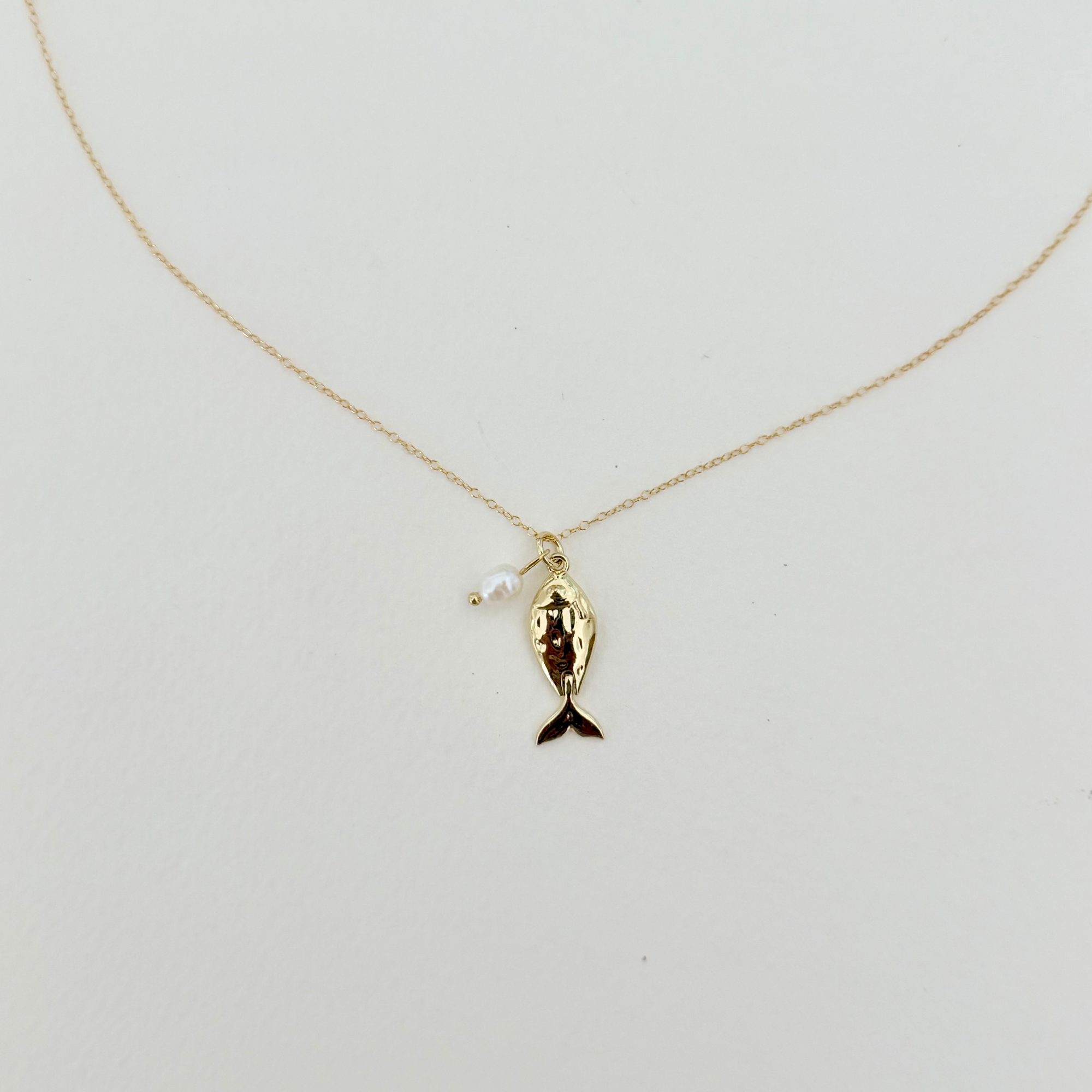 A delicate gold chain necklace featuring a small gold fish pendant with a dark tail and a tiny white pearl bead, displayed against a plain off-white background.