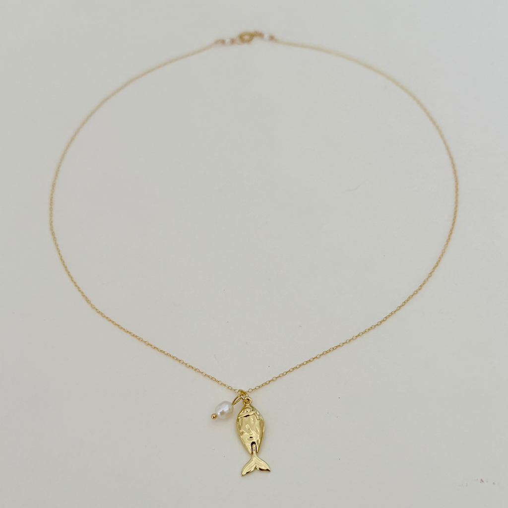 A delicate gold chain necklace featuring a small gold fish pendant and a tiny white pearl accent, displayed on a plain light background.