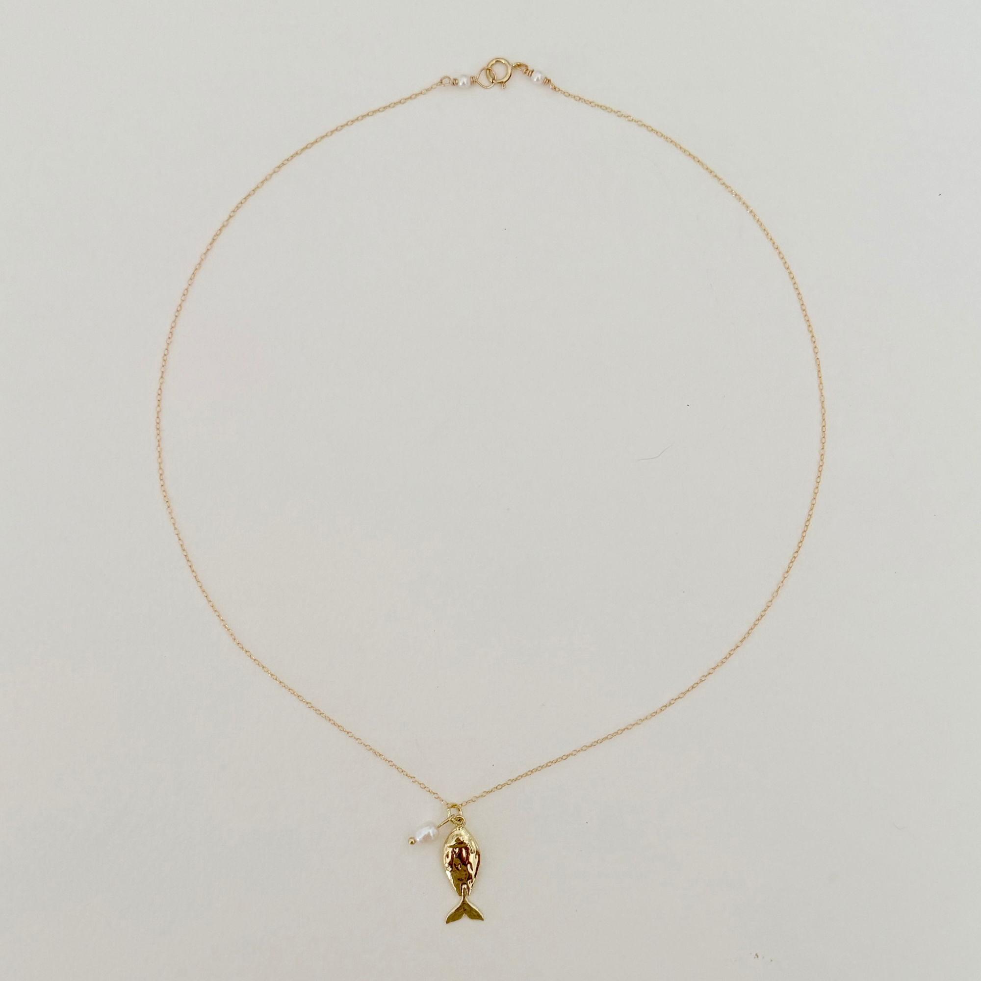 A delicate gold chain necklace featuring a small gold fish pendant with a red enamel face, displayed against a plain light gray background.