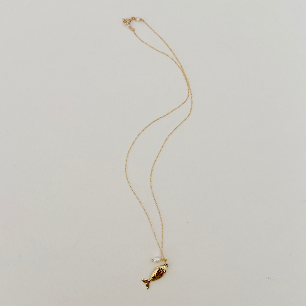 Delicate gold necklace featuring a fish pendant and pearl charm on a light grey background