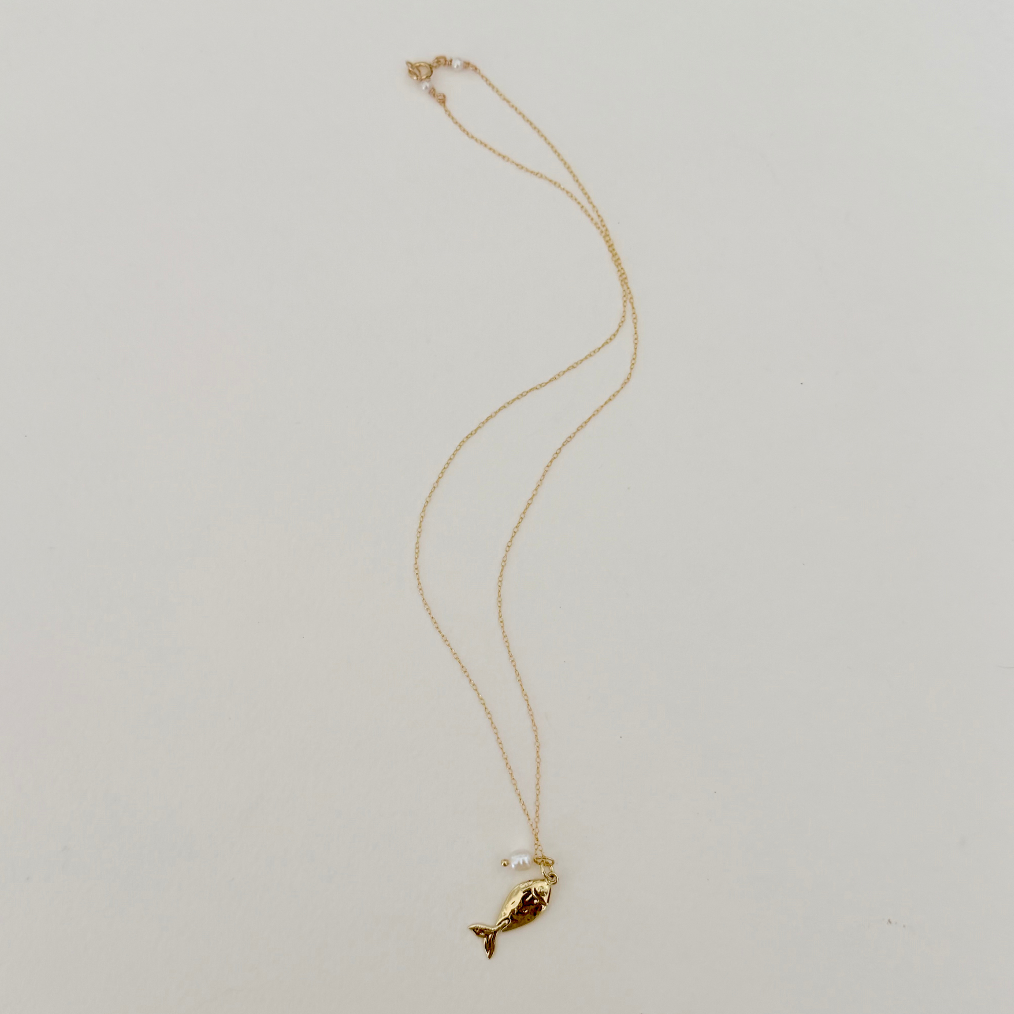 Delicate gold necklace featuring a fish pendant and pearl charm on a light grey background