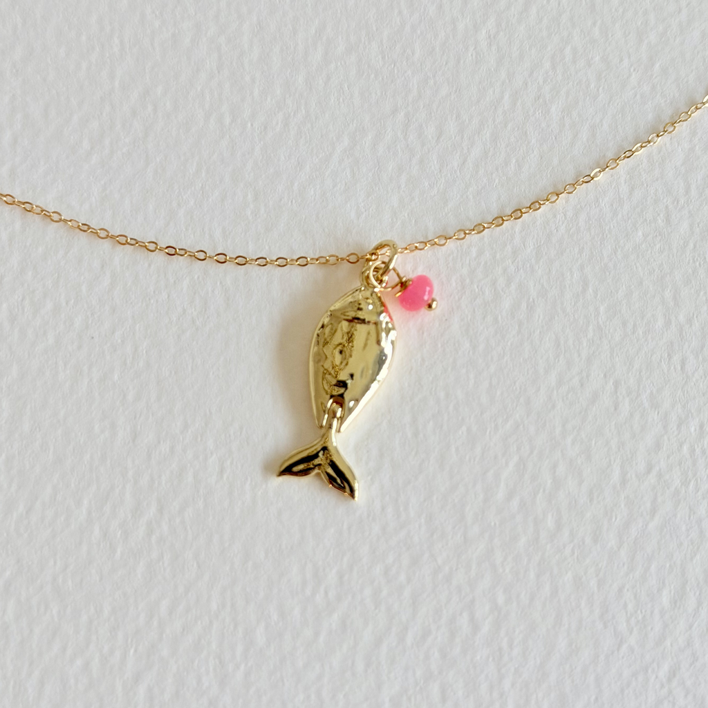 Gold fish-shaped pendant with a pink heart on a gold chain against a light gray background